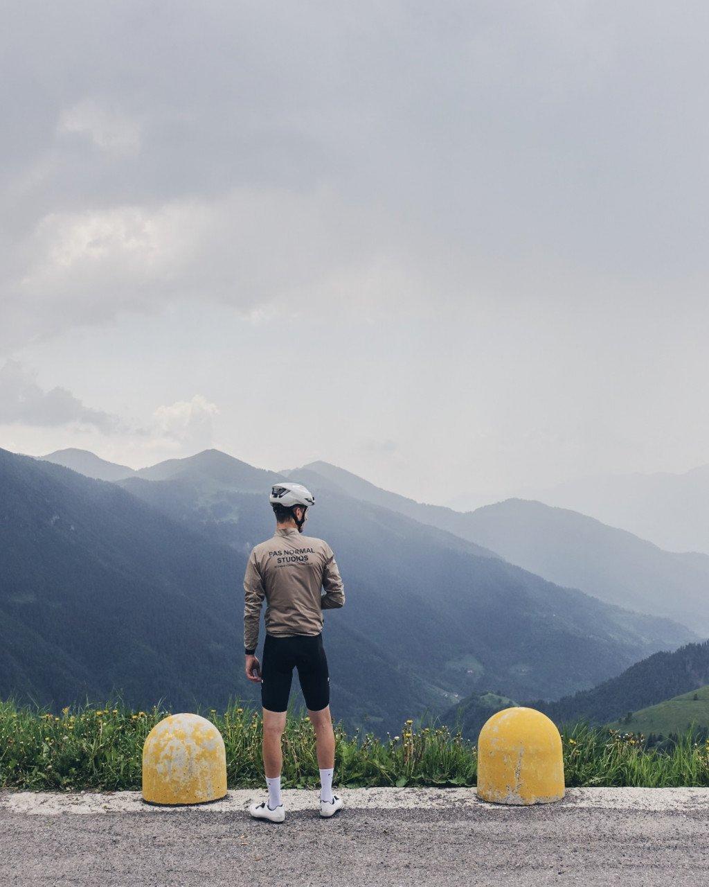 Model wearing beige long sleeve jersey with back branding and black shorts, overlooking mountain landscape