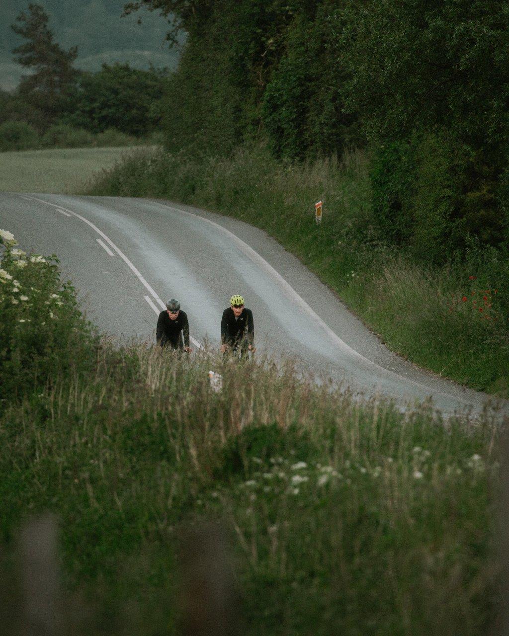 Two cyclists in dark cycling apparel riding on rural road through countryside