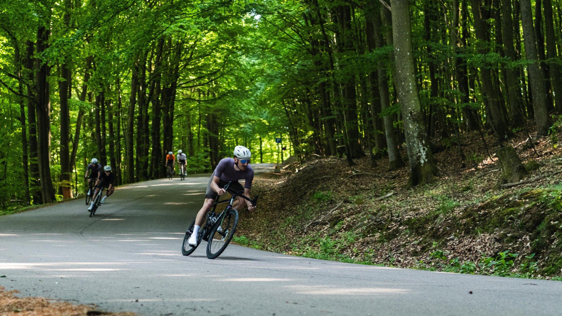 Cyclist in aerodynamic position on time trial bike riding through forested road with other cyclists in background