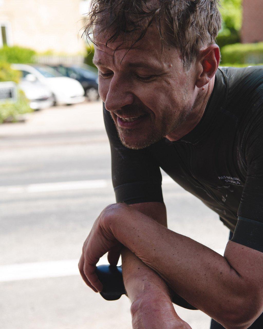 Model wearing black cycling jersey with chest logo, crouched position on street
