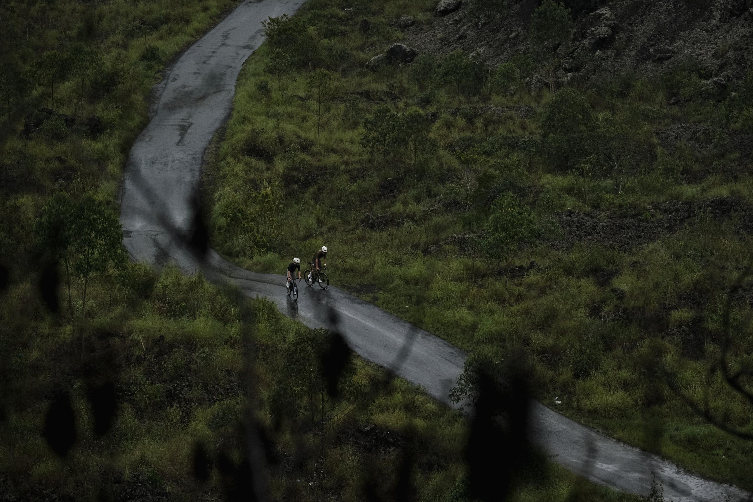 Two cyclists on winding mountain road through green landscape in Bali