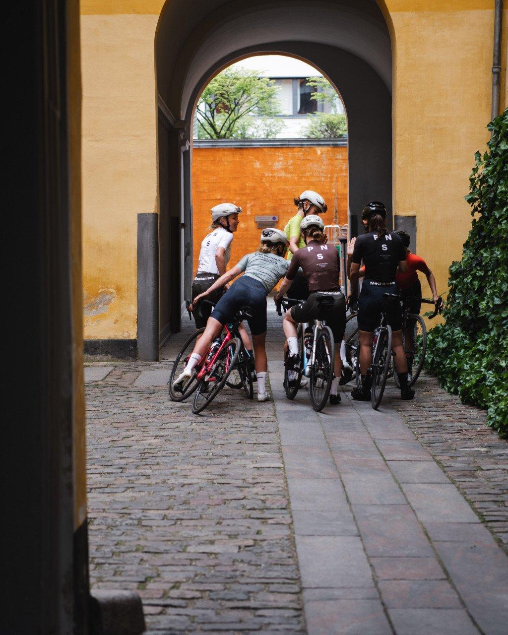 Cyclists in team jerseys gathering with bikes under stone archway on cobblestone street