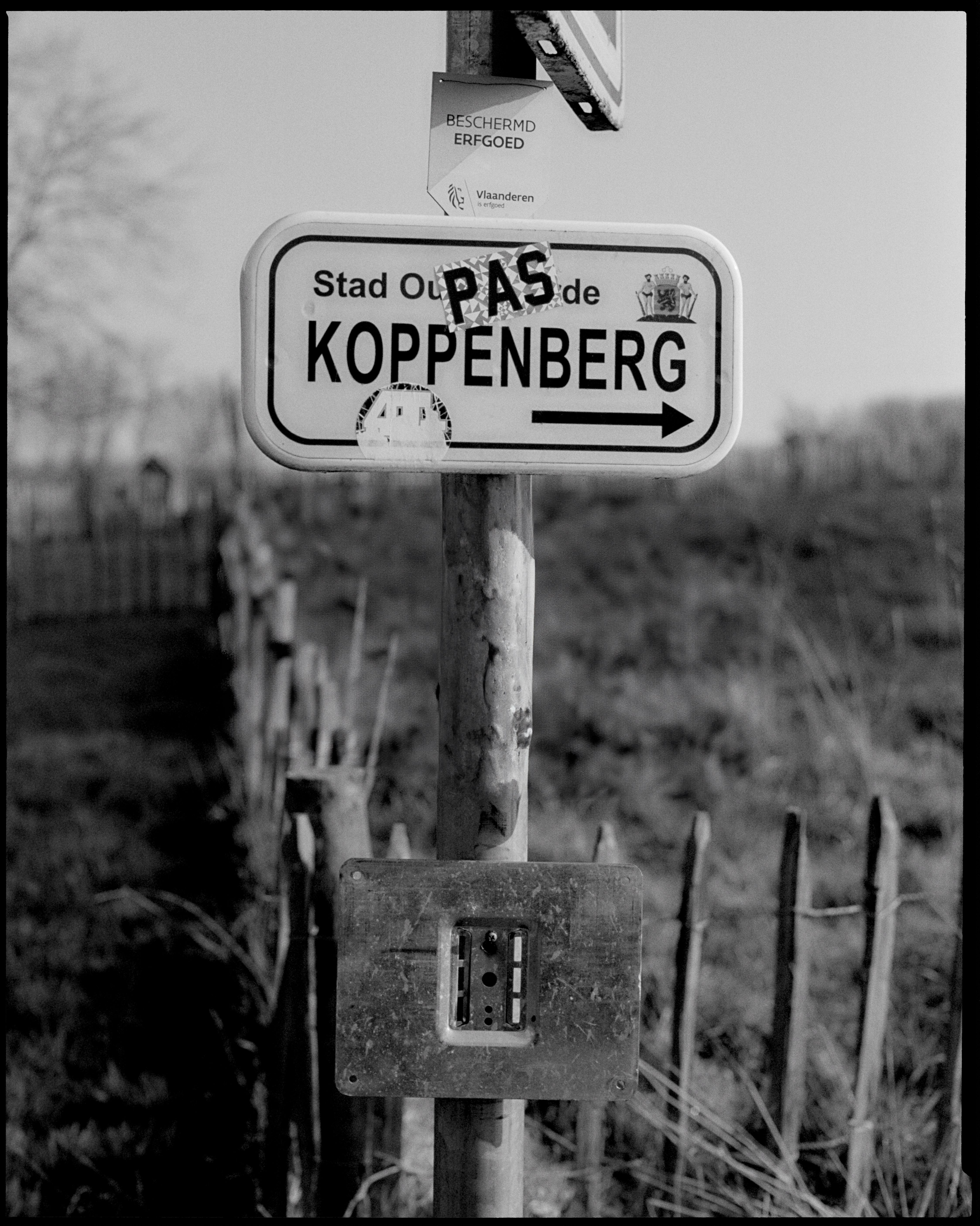 Black and white photo of Koppenberg road sign with PAS collection branding in Belgian countryside