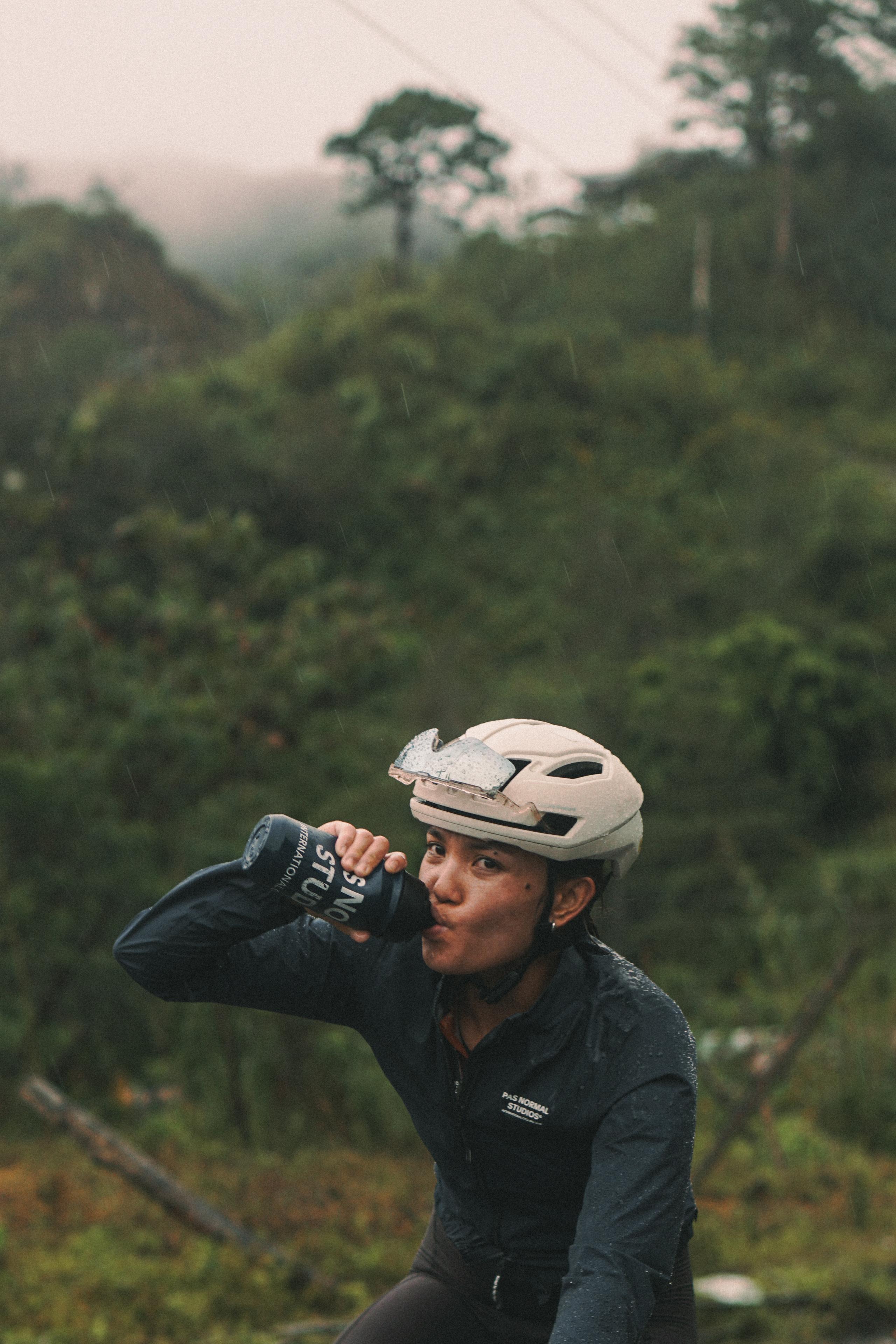 Model wearing beige cycling helmet drinking from black water bottle in natural outdoor setting