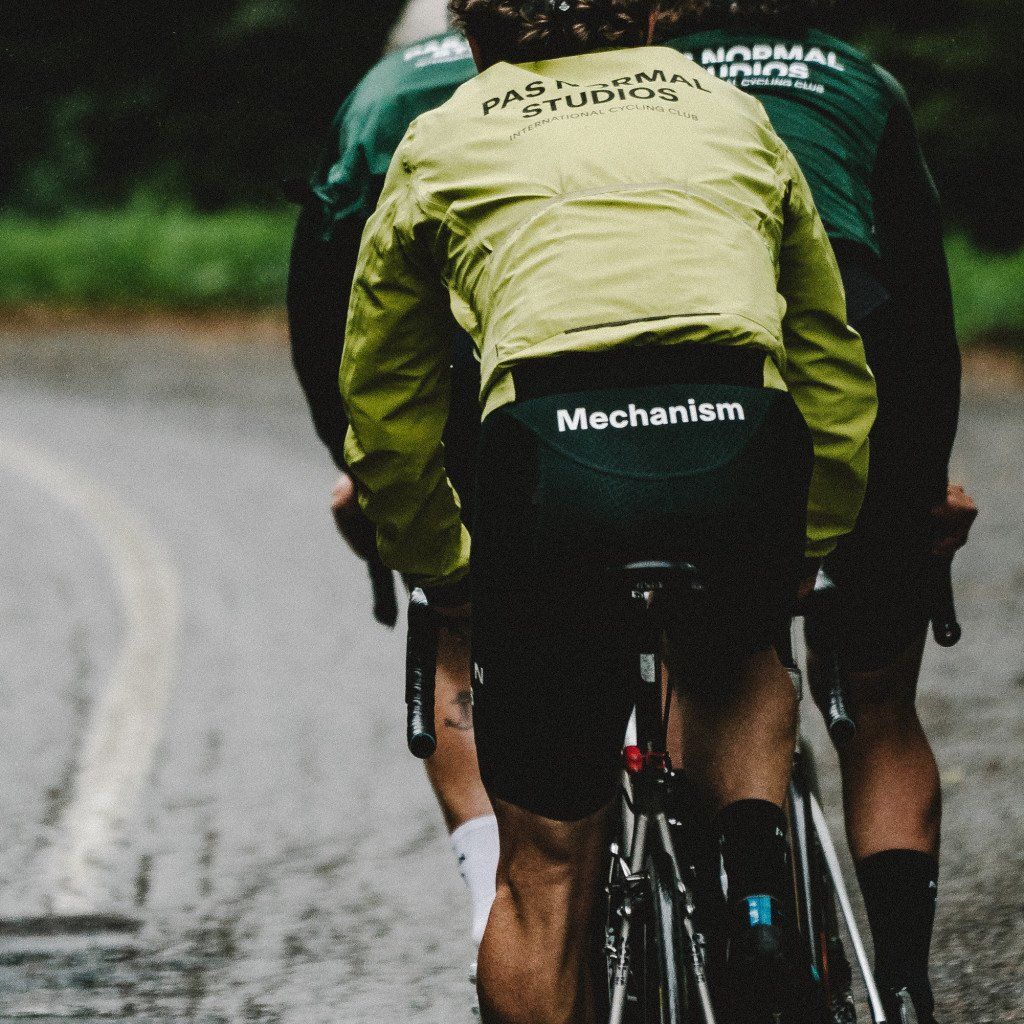 Model wearing Mechanism collection yellow and green jersey cycling on road, rear view