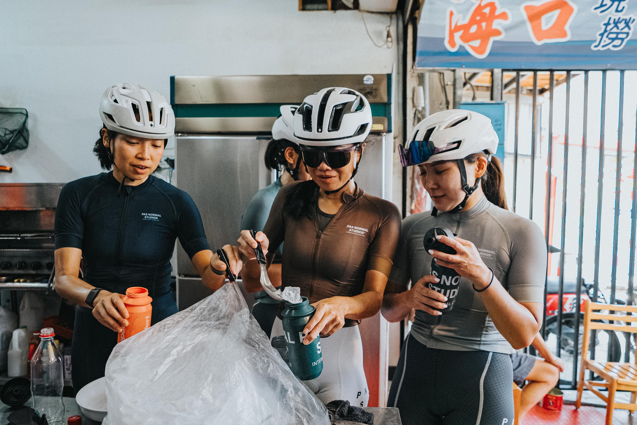 Three cyclists wearing white helmets and cycling jerseys at food stall, holding drinks and snacks