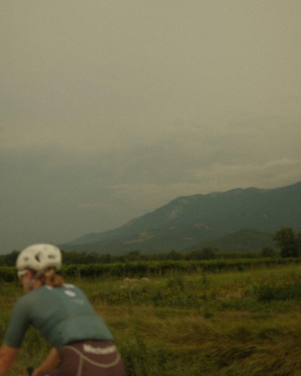 Cyclist in white helmet and light blue jersey riding through rural landscape with mountains in background