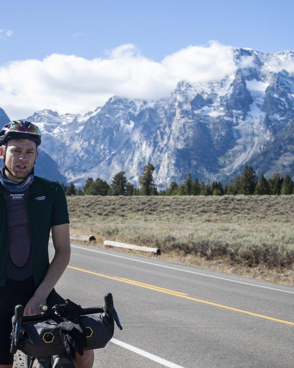 Model wearing dark green jersey with branding on mountain road with snow-capped peaks