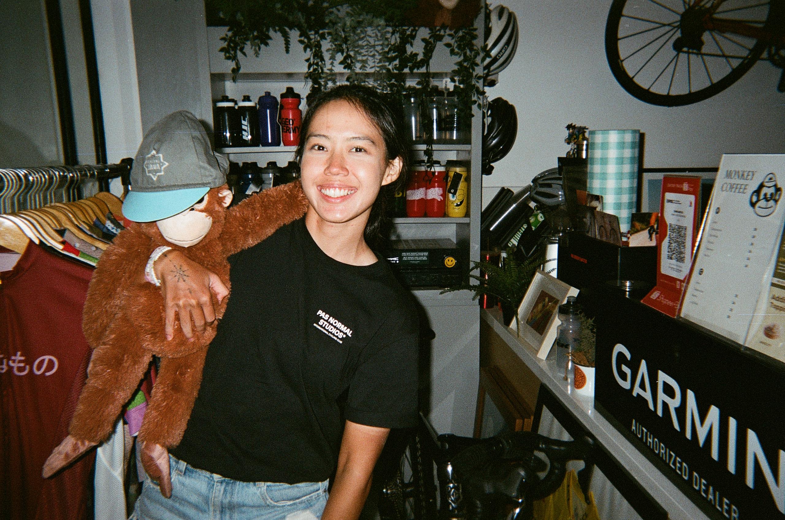Model wearing black short-sleeve t-shirt with white chest logo in bicycle shop setting