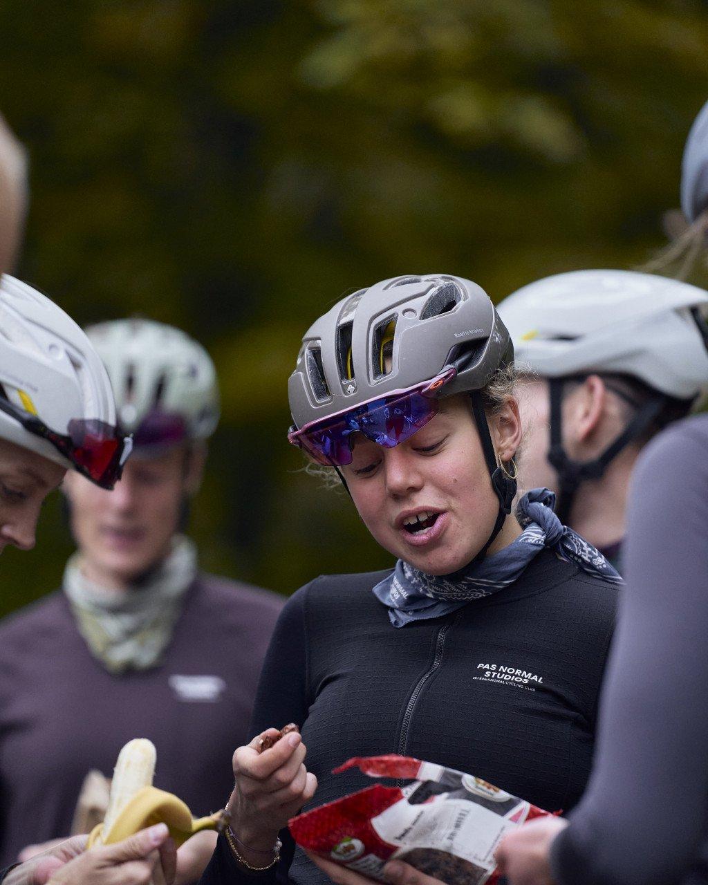 Model wearing black cycling jacket with white branding, white helmet, and blue sunglasses during group ride