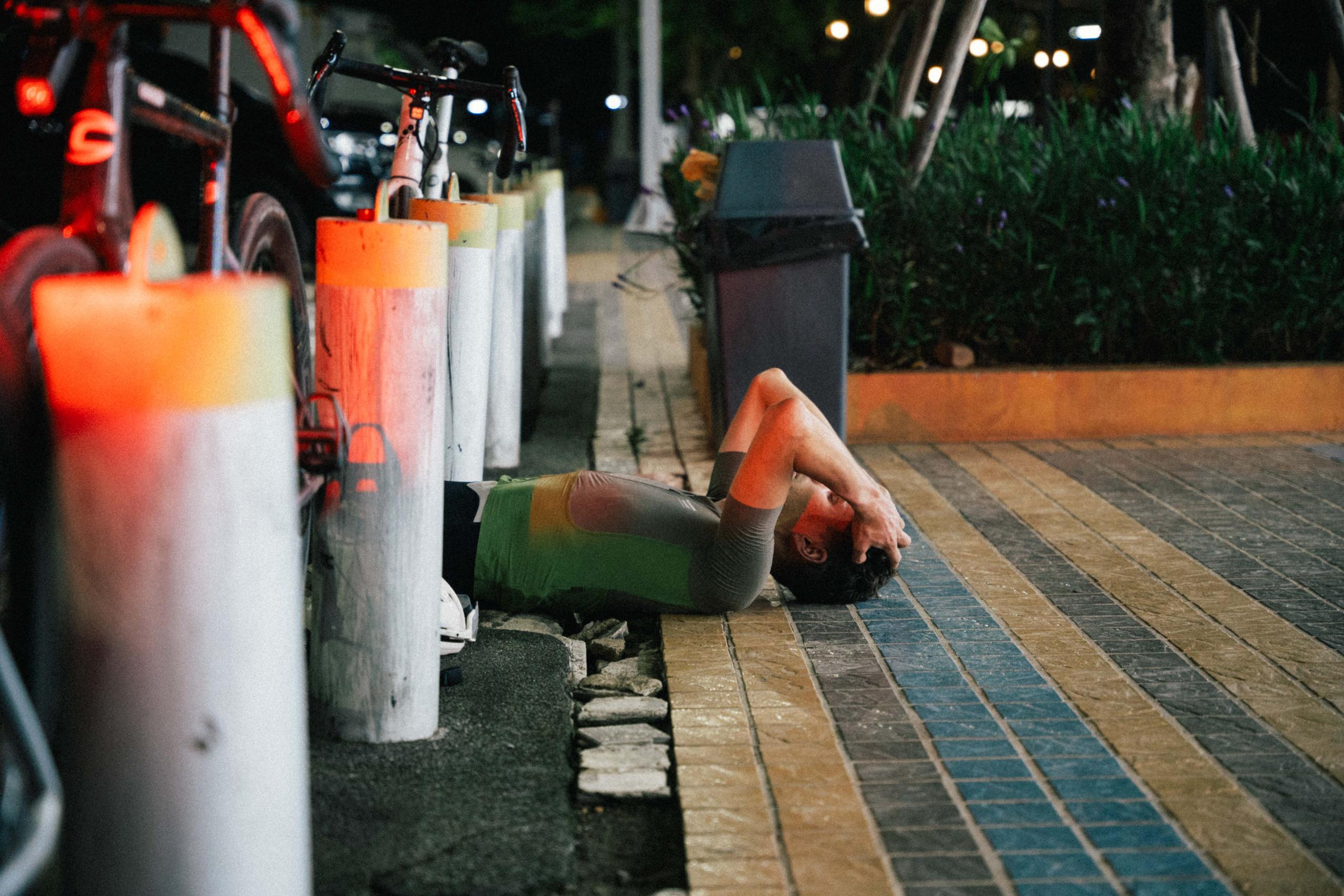 Model wearing orange jersey and green cycling shorts resting on pavement beside bike in urban nighttime setting