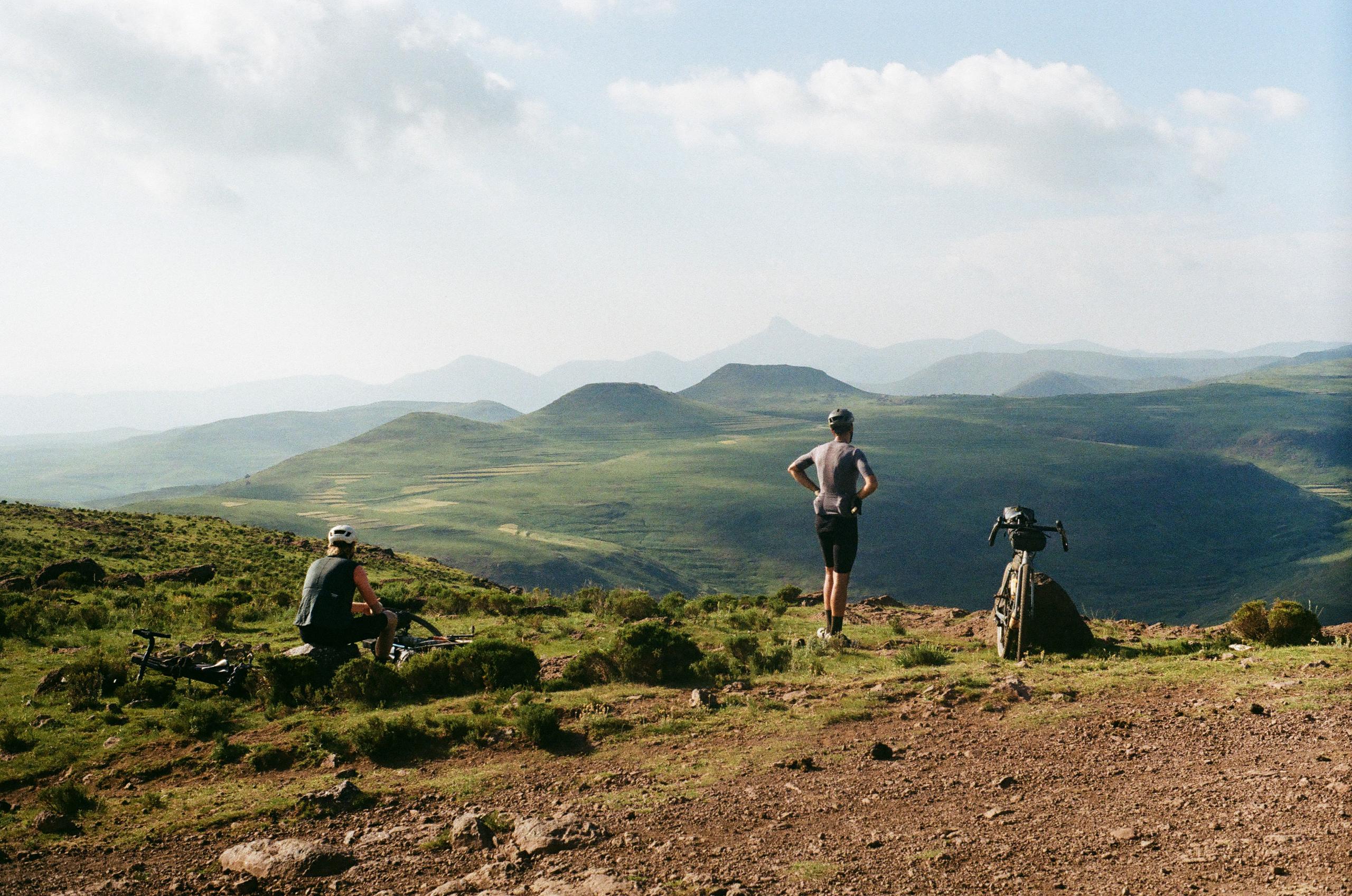 Two cyclists taking break on mountain terrain with rolling hills backdrop in Lesotho