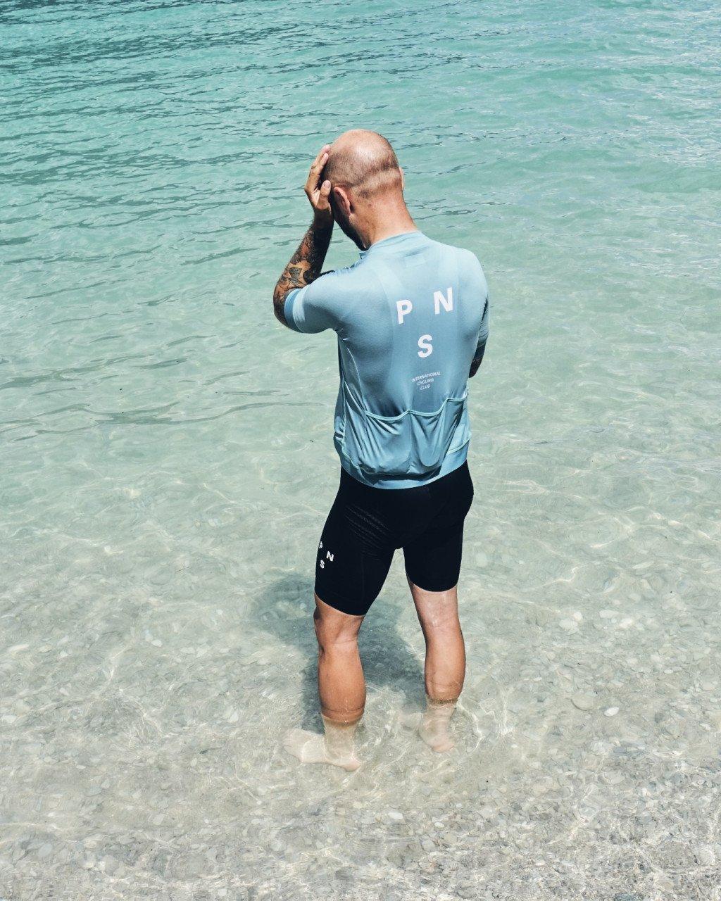 Model wearing light blue long sleeve jersey with back logo and black shorts standing in clear ocean water