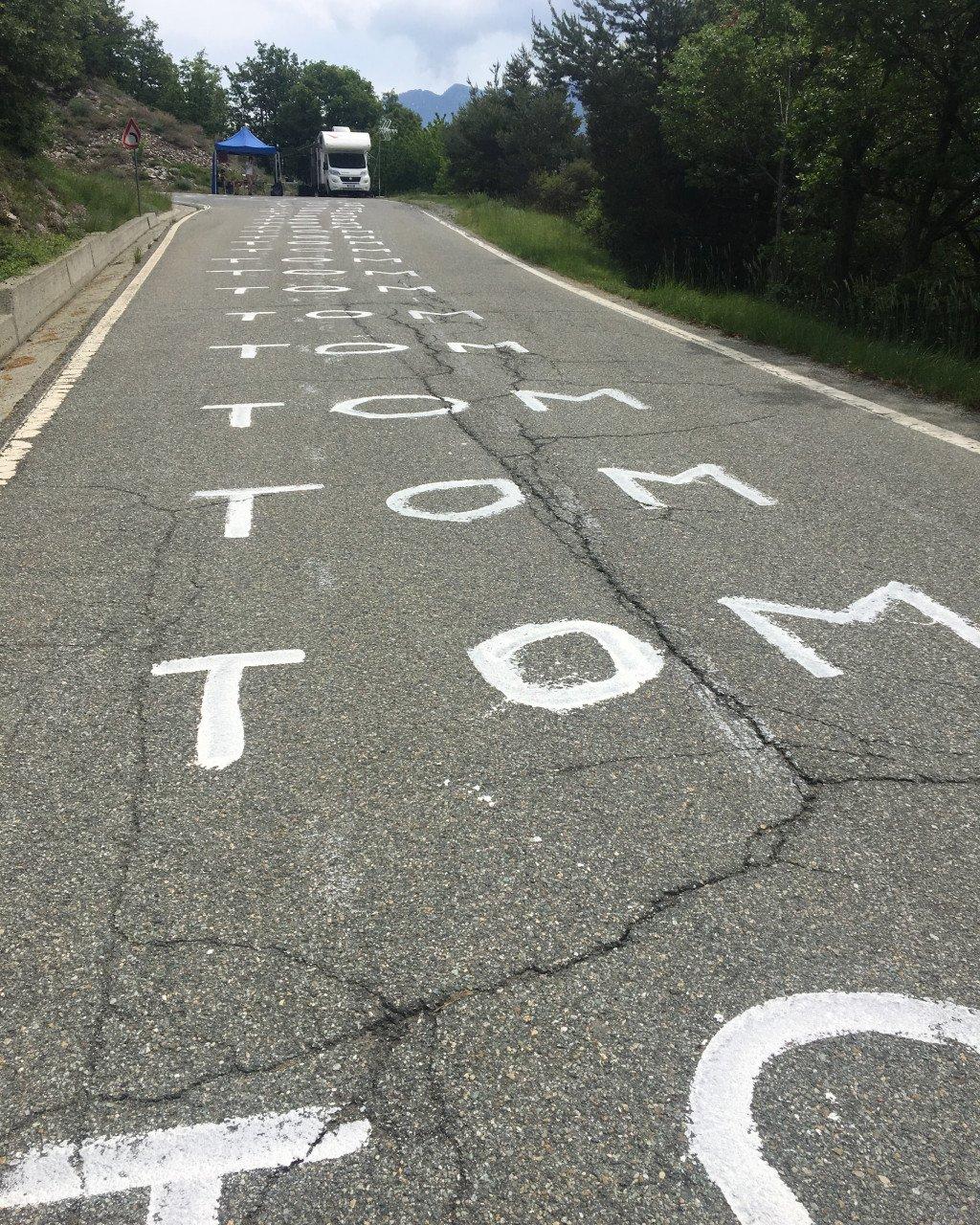 Road with white painted letters spelling Tour de France stage markers in mountainous countryside