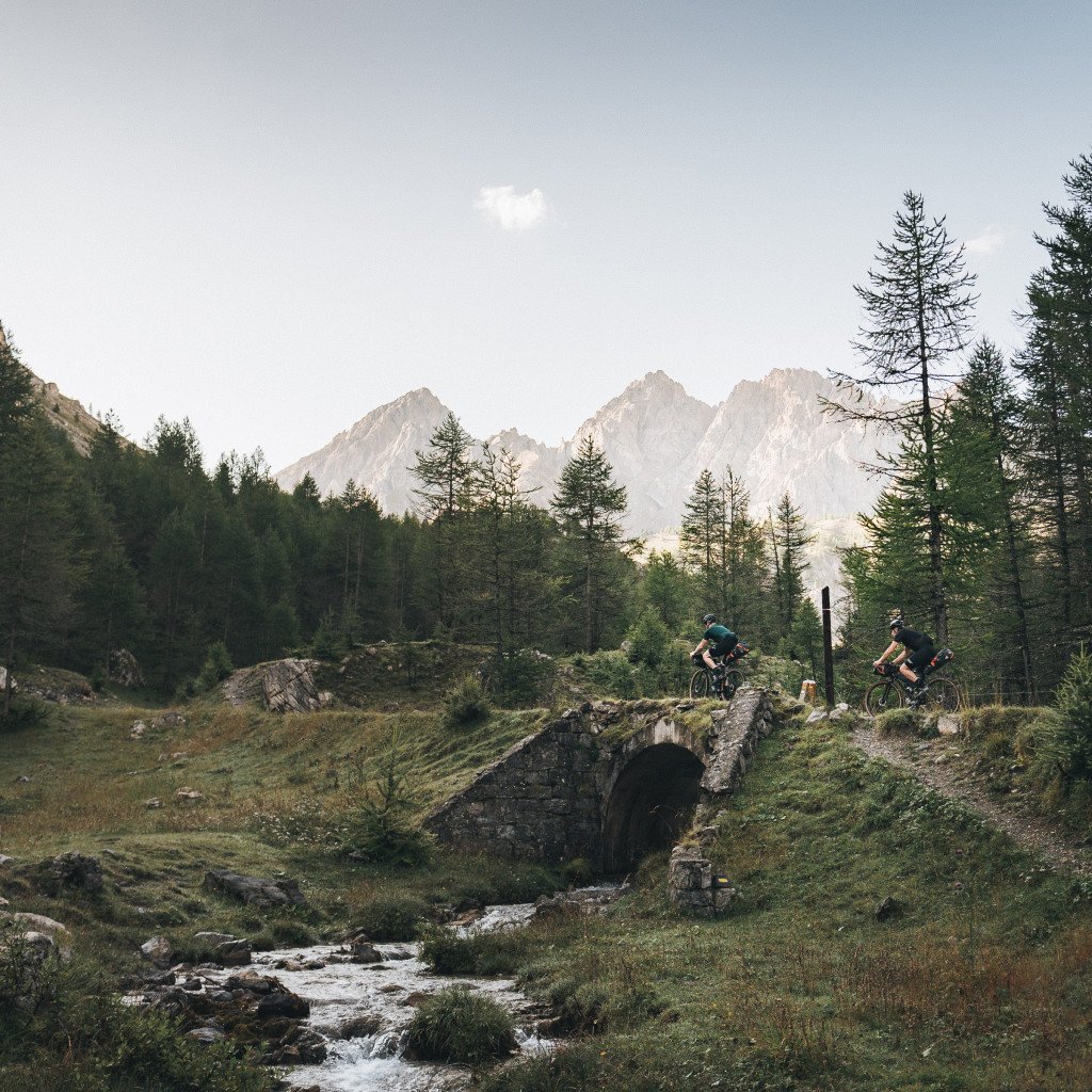 Two cyclists crossing stone bridge over mountain stream with alpine peaks and forest backdrop