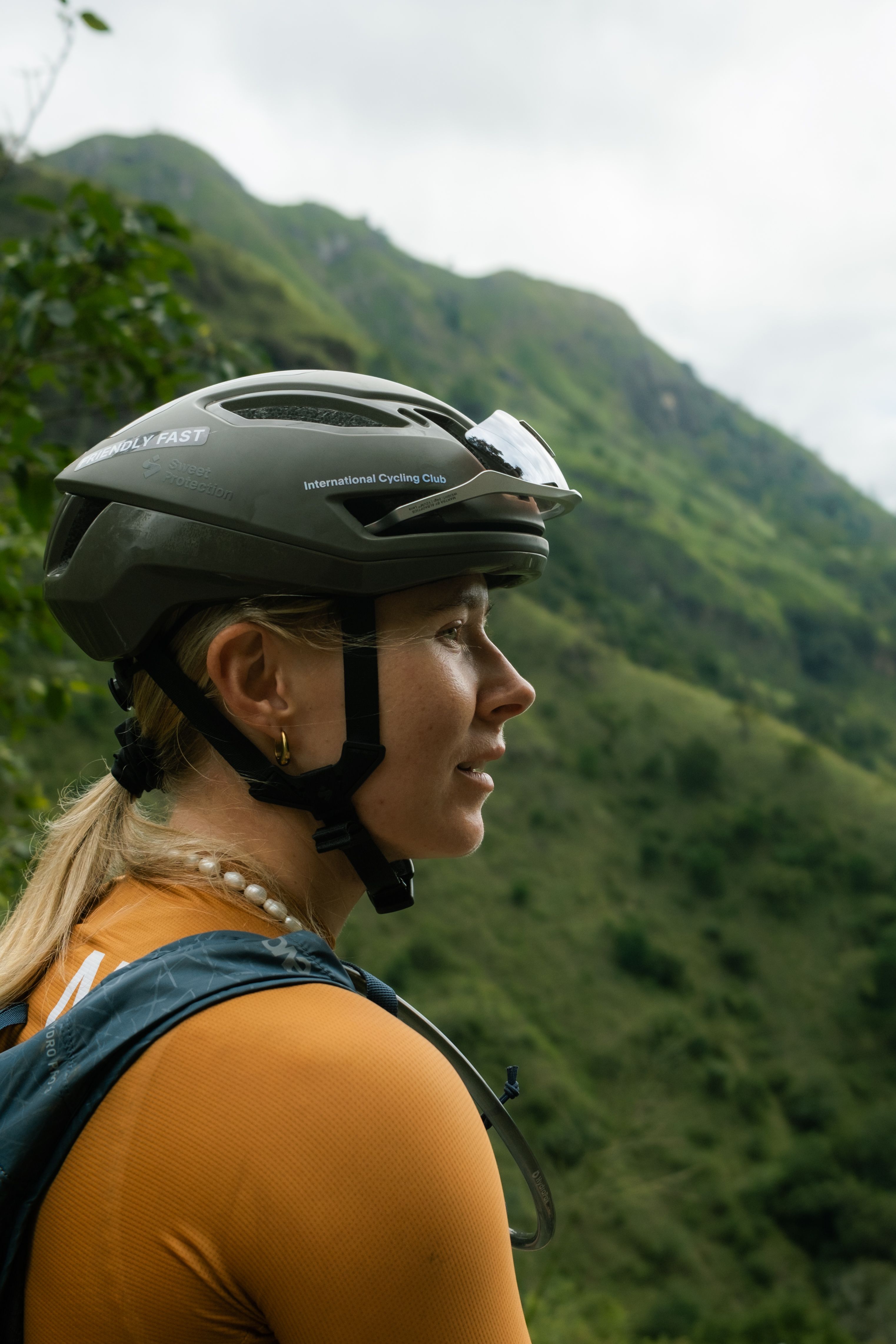 Female cyclist wearing orange jersey and olive green cycling helmet in mountainous landscape