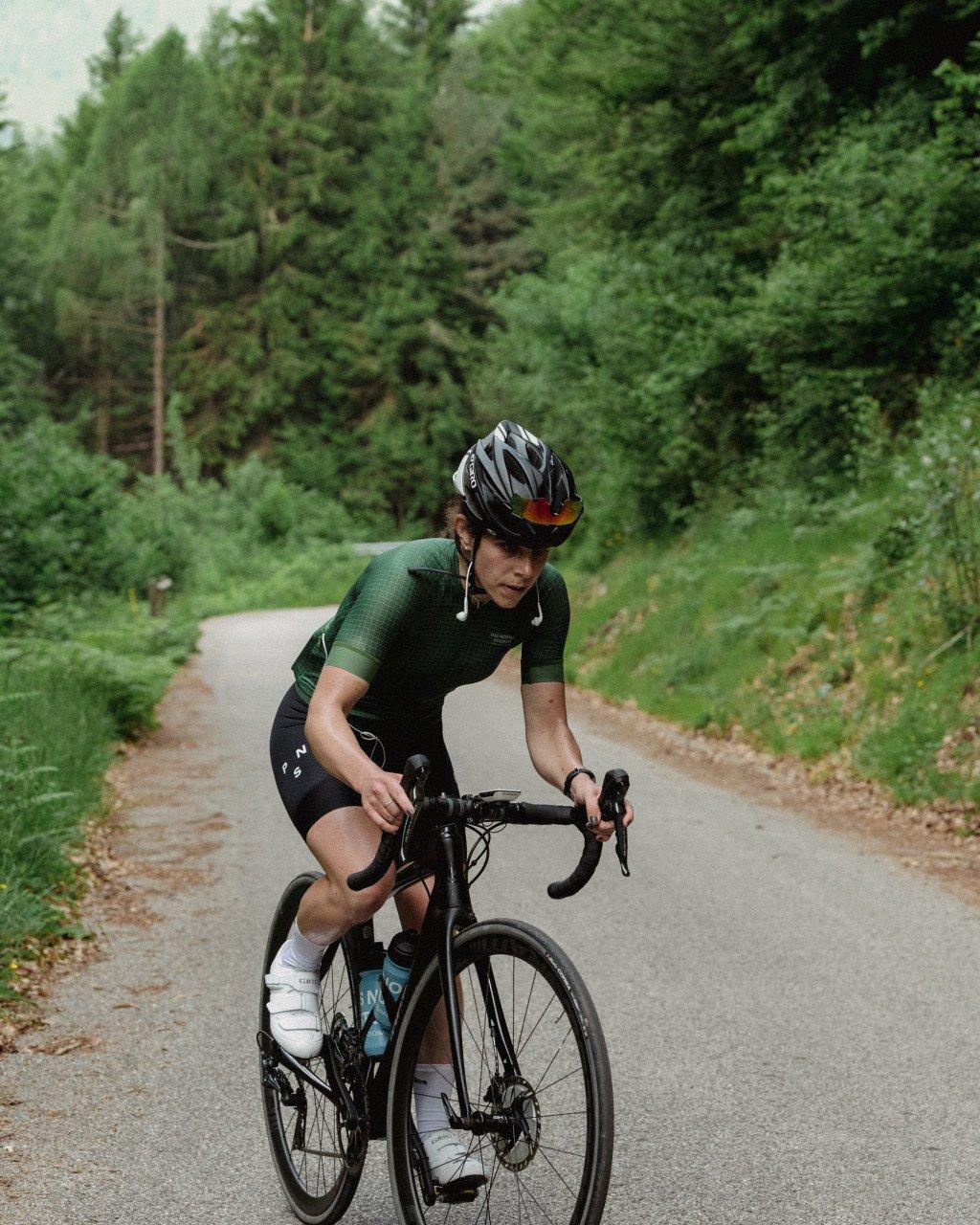 Model wearing green cycling jersey and black shorts cycling on country road through forest