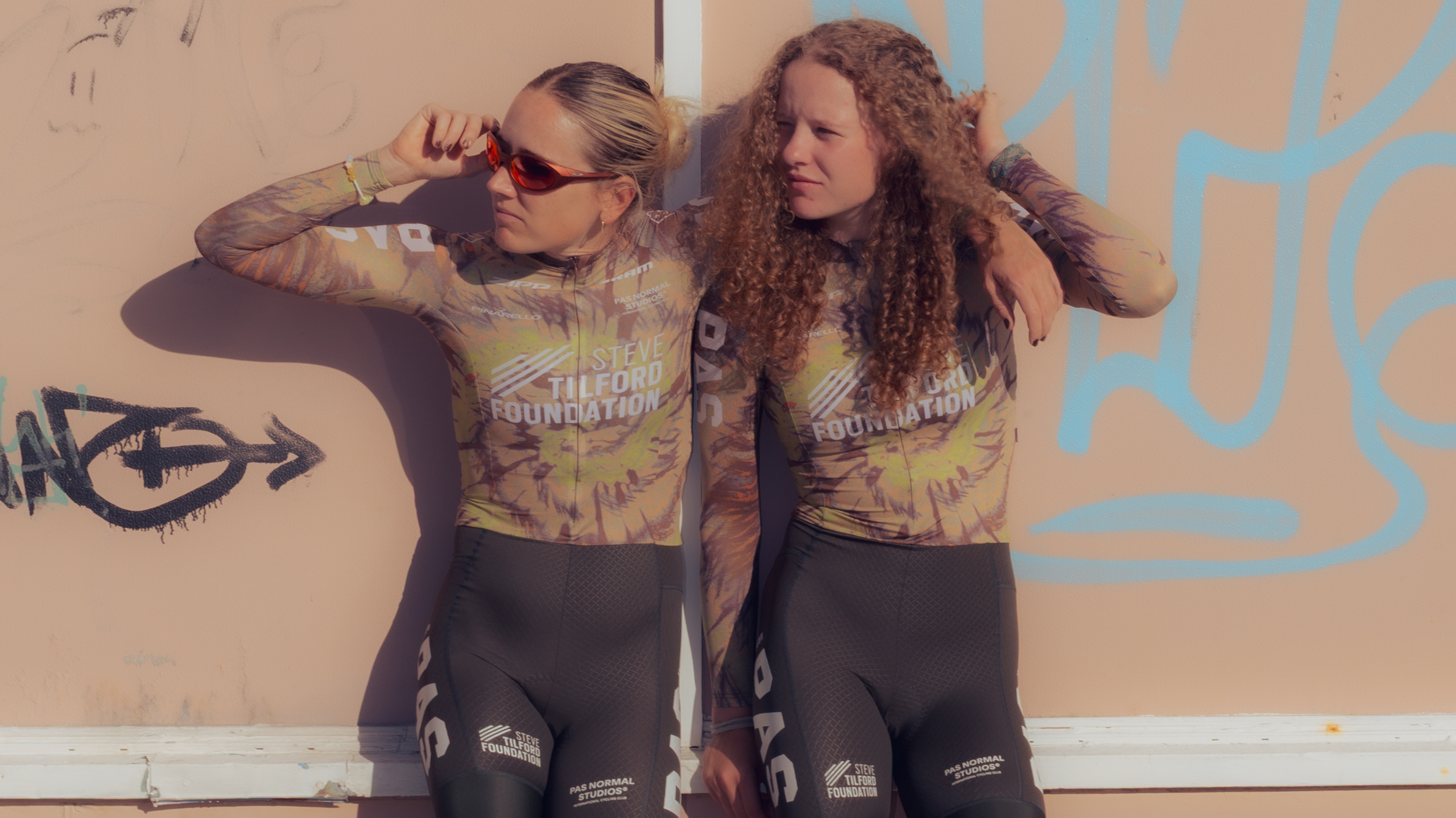 Models wearing Steve Tilford Foundation racing kit with tie-dye pattern and black shorts against graffiti wall