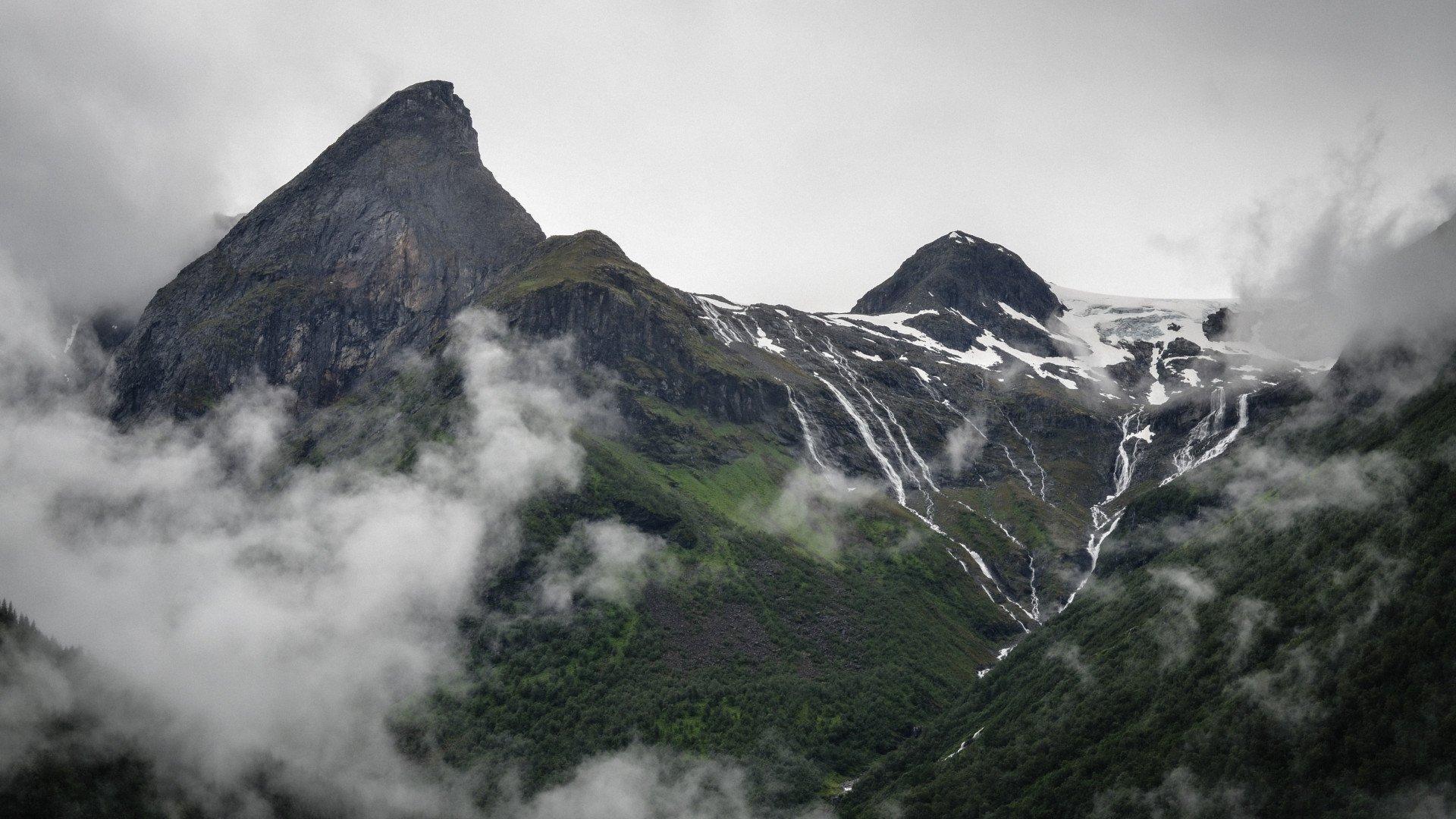 Dramatic Norwegian mountain landscape with snow-capped peaks, waterfalls, and misty clouds over green valleys