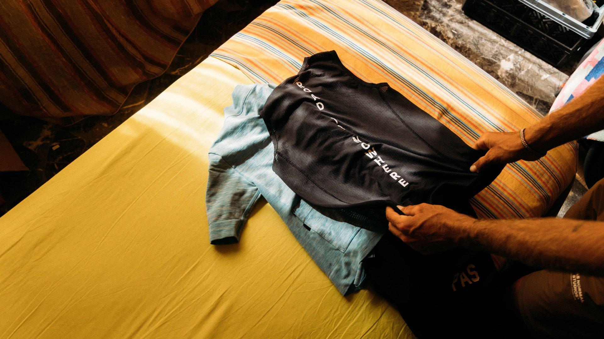 Model examining black cycling jacket with chest lettering alongside tie-dye garment on yellow surface