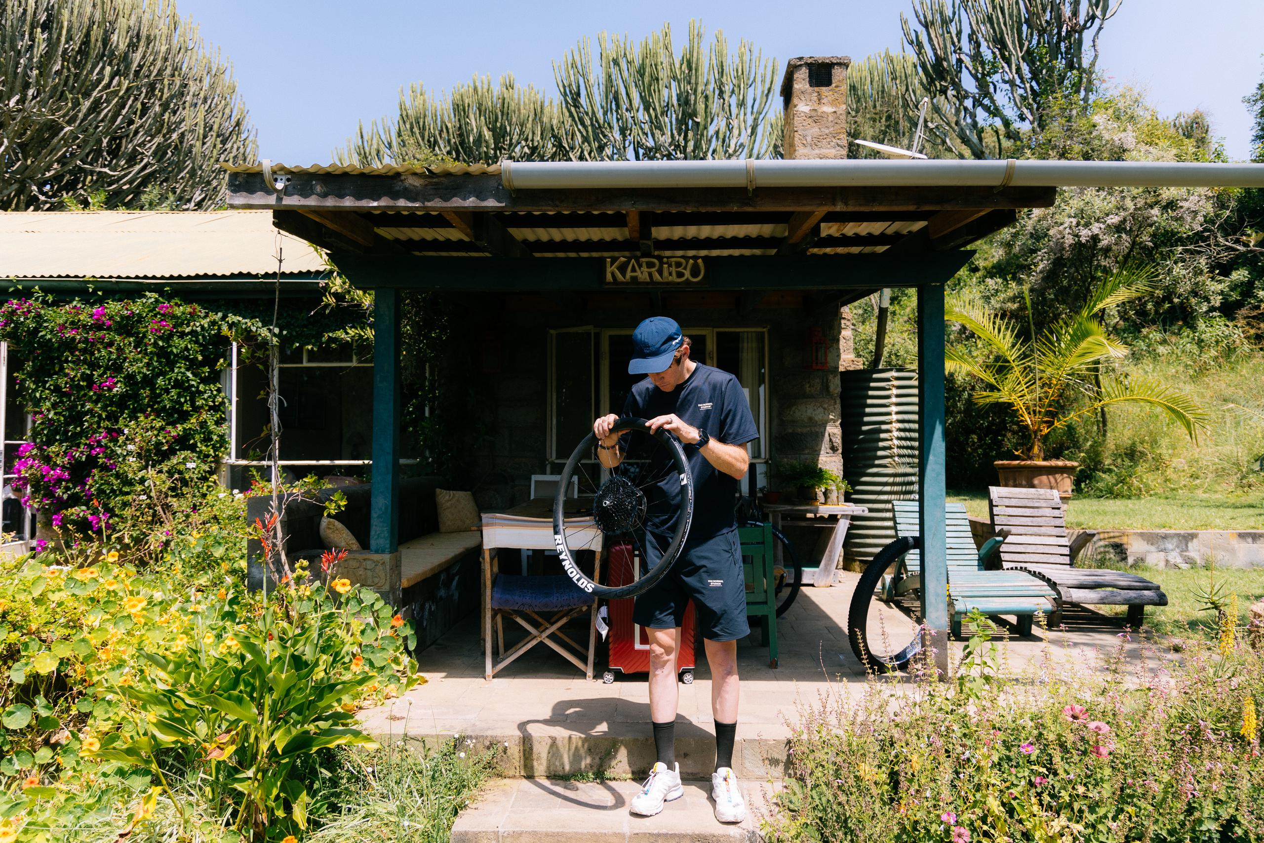 Model wearing navy t-shirt and shorts with blue cap, cleaning bicycle wheel at tropical rest stop