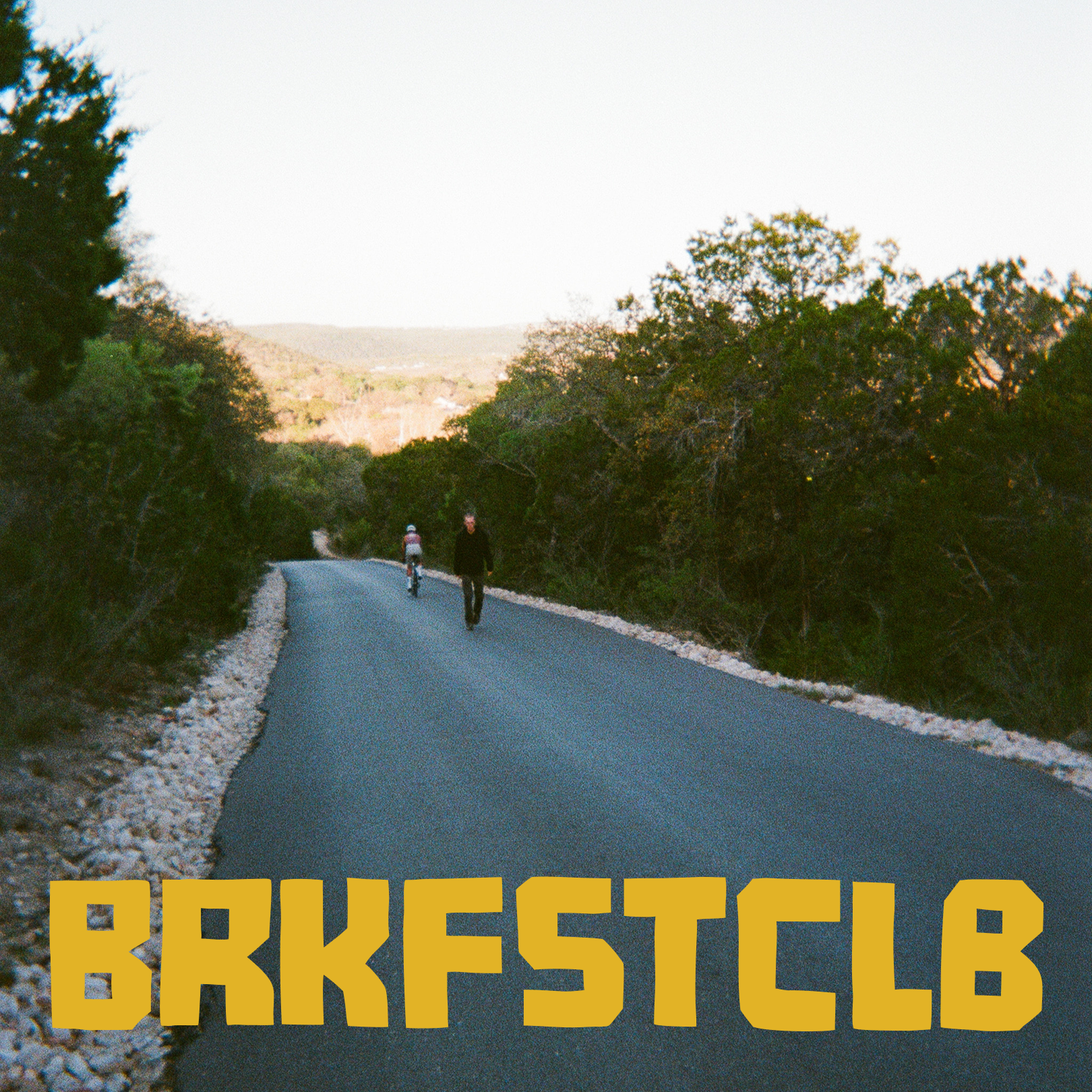 Cyclists riding on tree-lined road with BRKFSTCLB text overlay