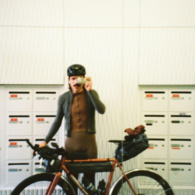 Model wearing dark cycling kit adjusting sunglasses next to orange bike in urban setting with white storage lockers