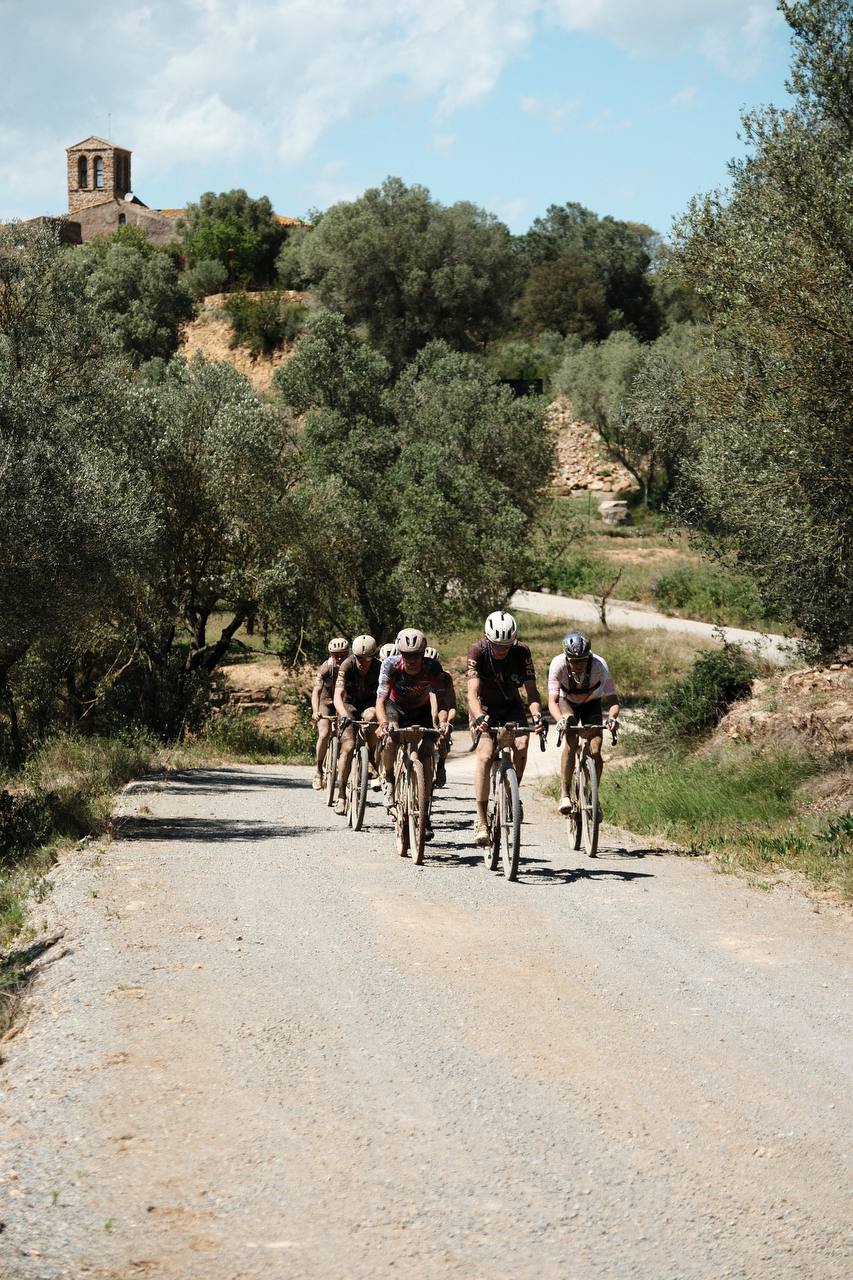 Cyclists in aerodynamic position riding time trial bikes on rural gravel road through Mediterranean landscape