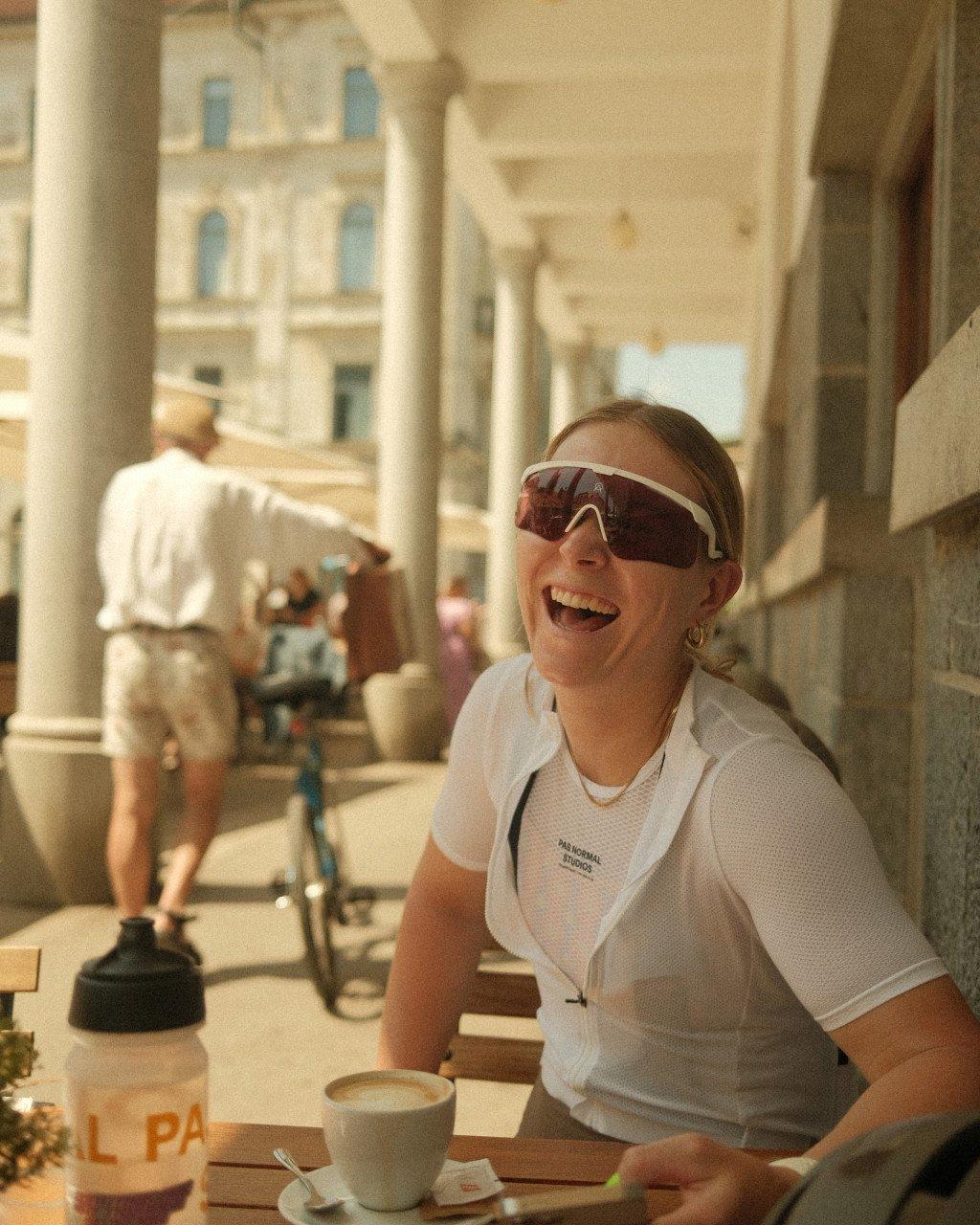 Model wearing white cycling jersey with branding and sunglasses, sitting at cafe table