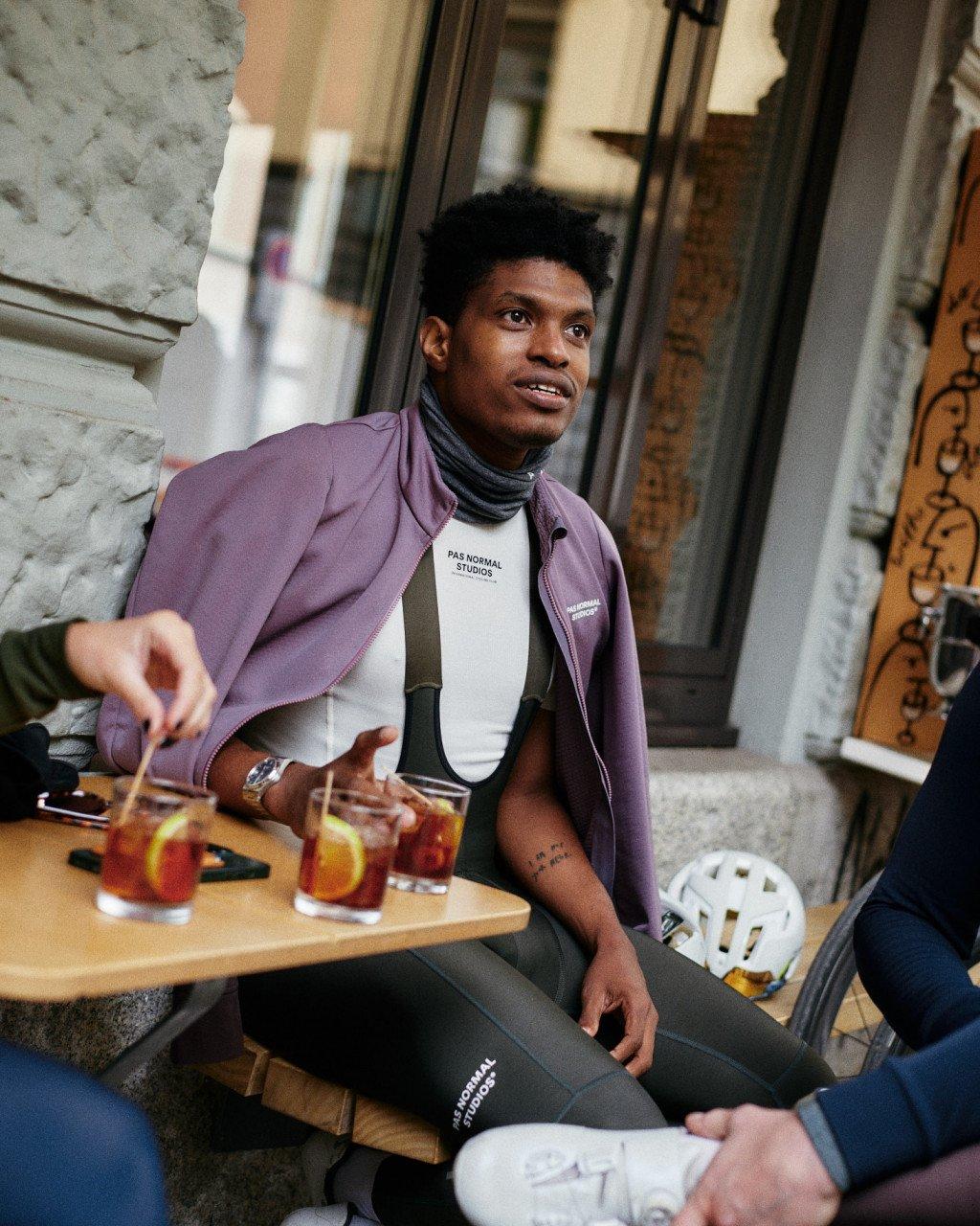 Model wearing purple jacket over white logo tee and dark tights, sitting at cafe table in urban setting