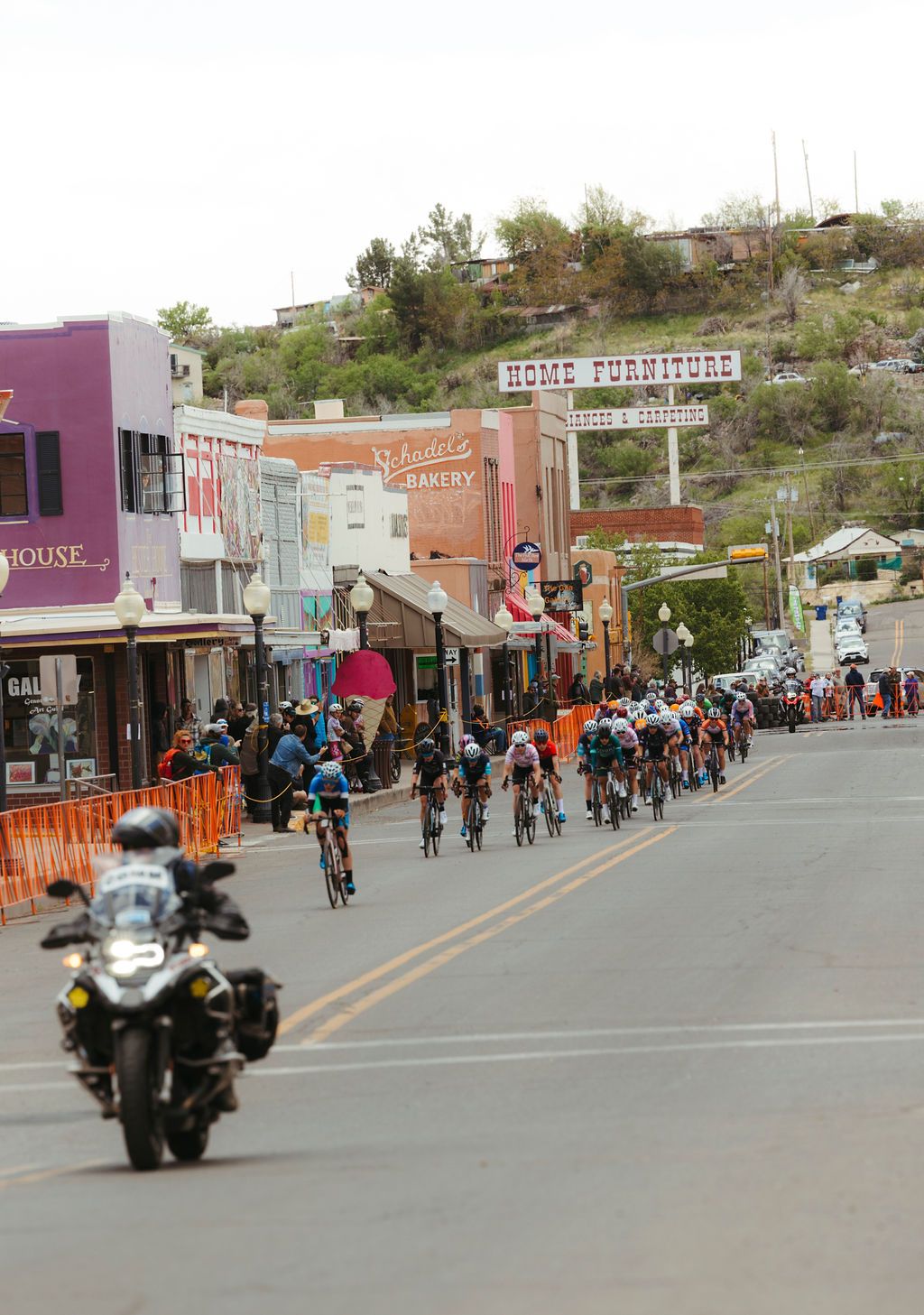 Cyclists racing through small town main street during Tour of the Gila with spectators and orange barriers