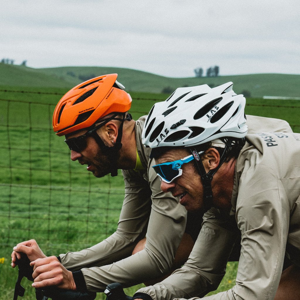 Two cyclists in aerodynamic position wearing beige kit, one with orange helmet and one with white helmet, racing through countryside