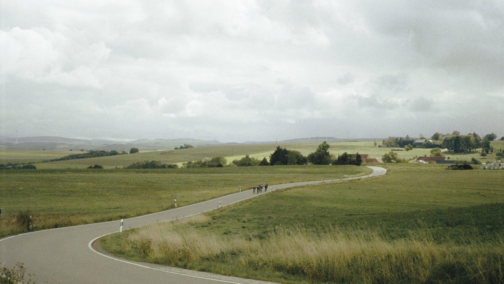 Cyclists on winding country road through rolling green countryside under cloudy sky