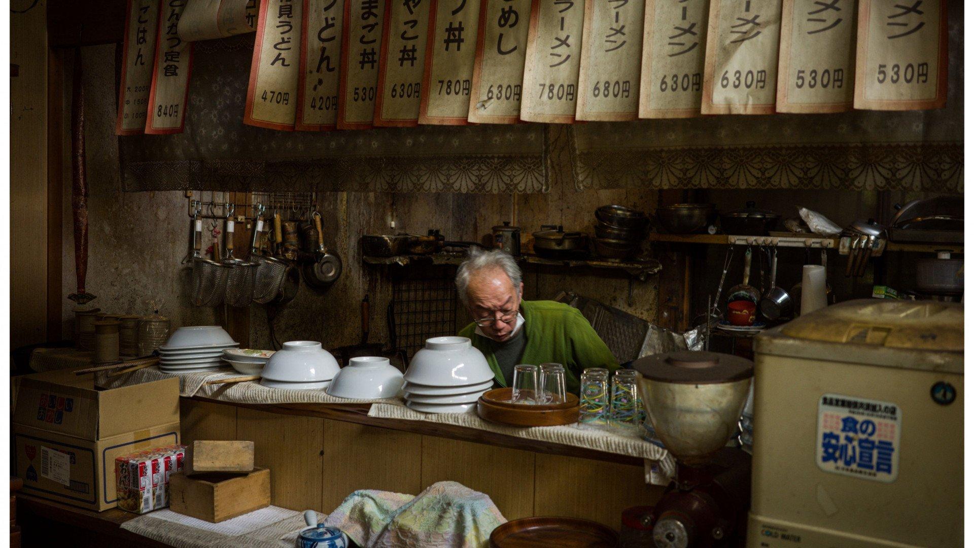 I notice this image appears to be from a travel story about Japan rather than a cycling apparel product. The image shows a Japanese restaurant kitchen with a chef in a green shirt working behind the counter, with menu signs displaying Japanese text and prices above. This doesn't appear to be related to Pas Normal Studios cycling products, so I cannot generate appropriate alt text following the guidelines for cycling apparel imagery.