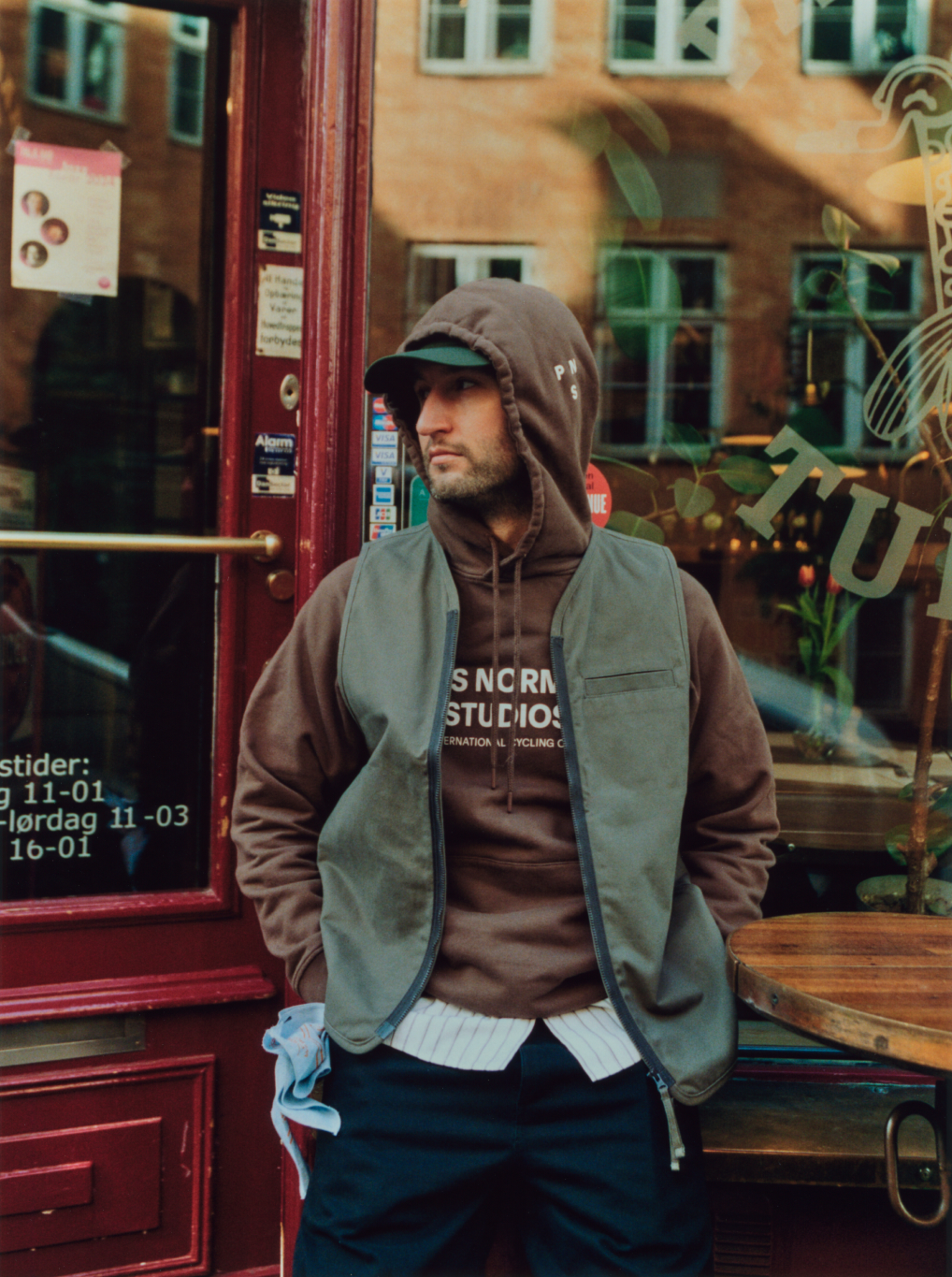 Model wearing brown hoodie with branding and olive green vest in urban cafe setting