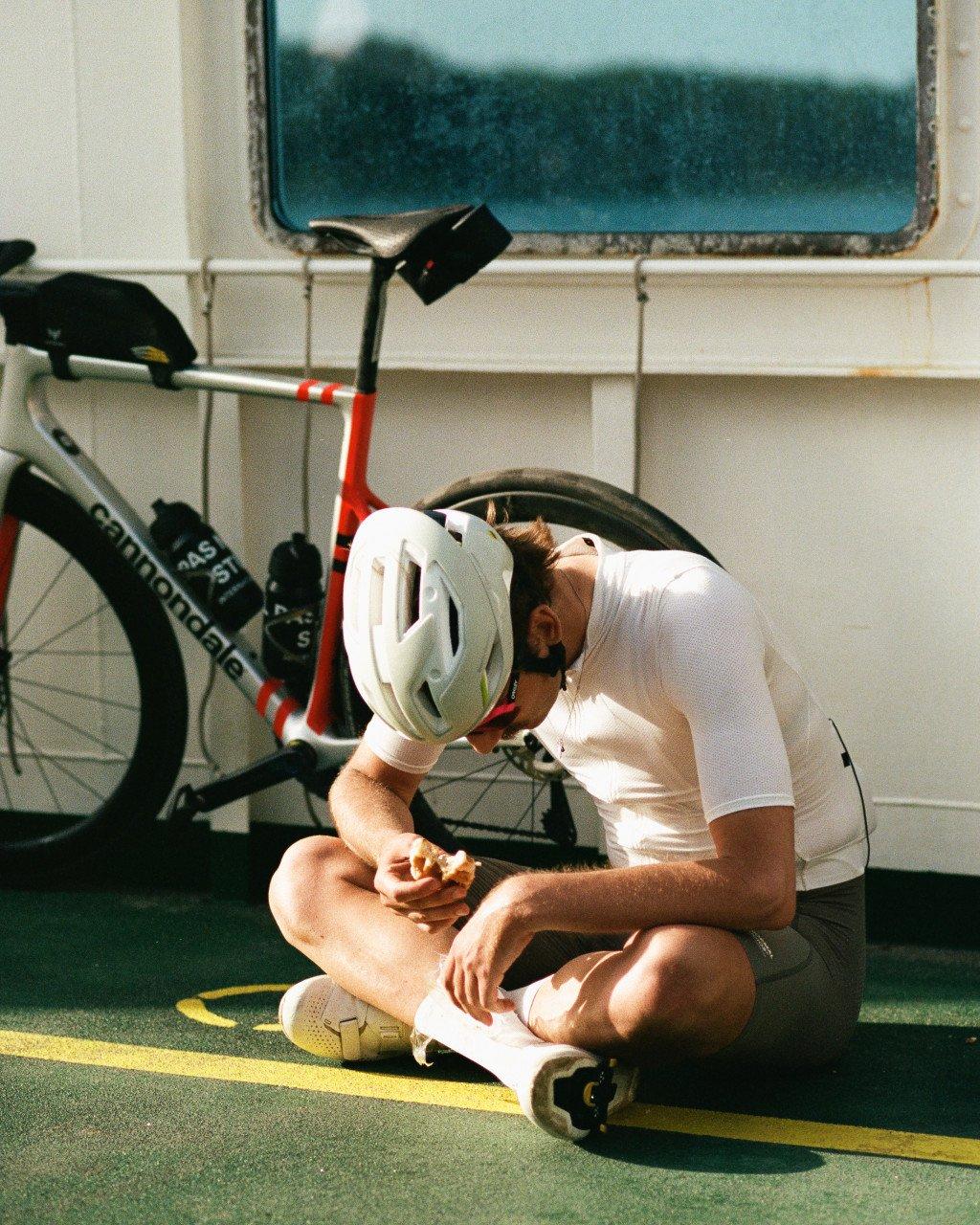 Model wearing white cycling jersey and shorts sitting on ferry deck with bicycle and helmet
