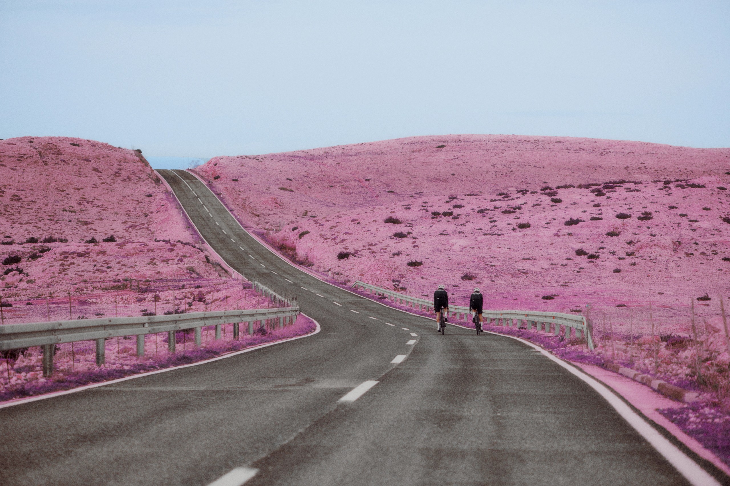 Two cyclists on winding desert road with pink-tinted landscape and rolling hills
