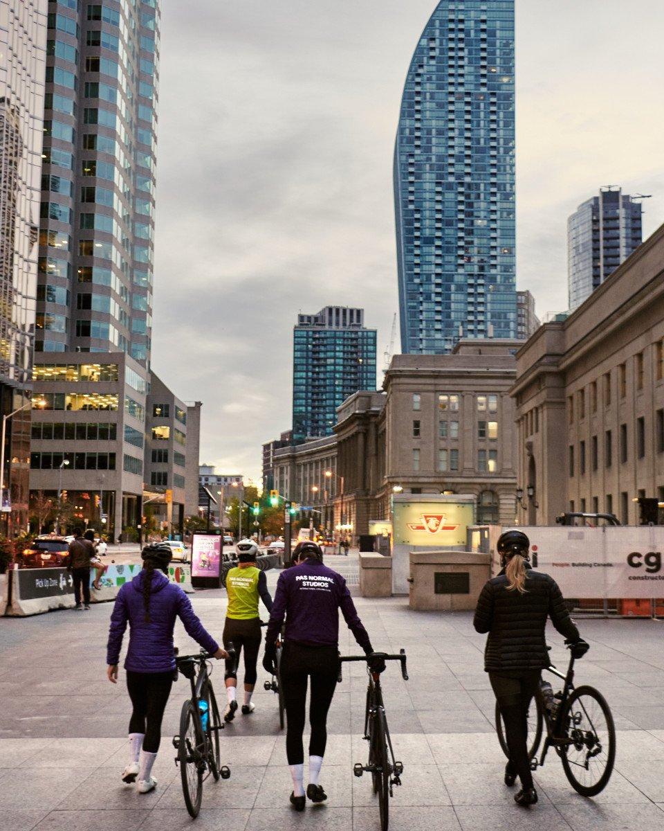 Cyclists walking with bikes through urban plaza at dusk, wearing purple and green cycling apparel