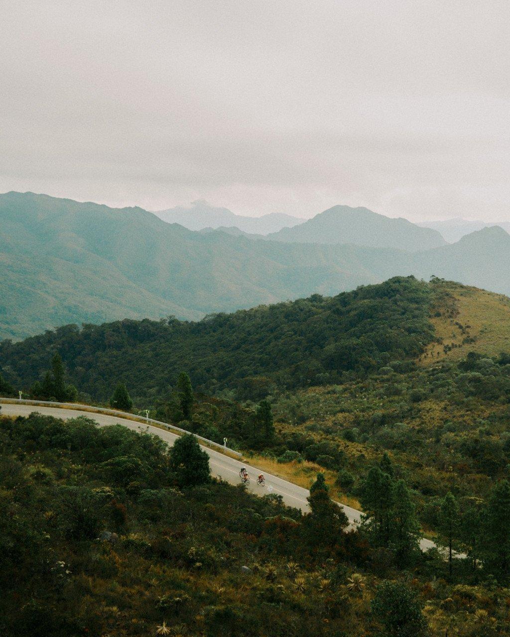 Two cyclists on winding mountain road through Colombian hills with layered mountain ranges in background