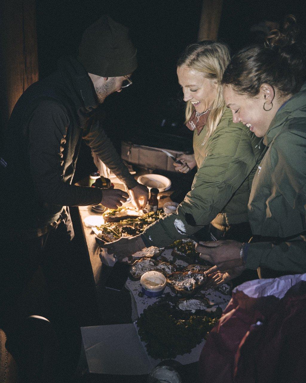 Group of cyclists in outdoor apparel gathered around campfire cooking food during evening camping trip