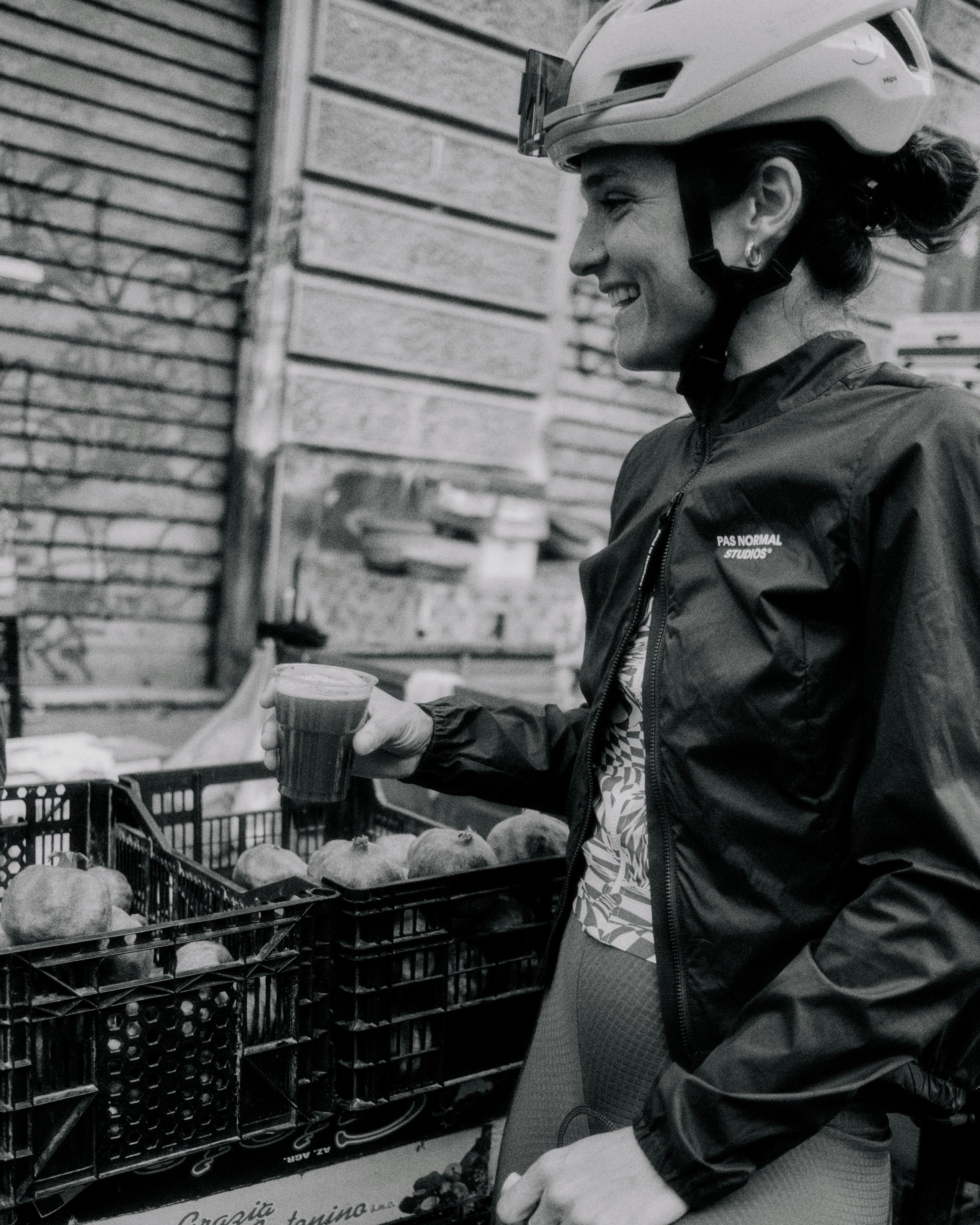 Model wearing dark cycling jacket with helmet and branding, holding coffee cup at outdoor market