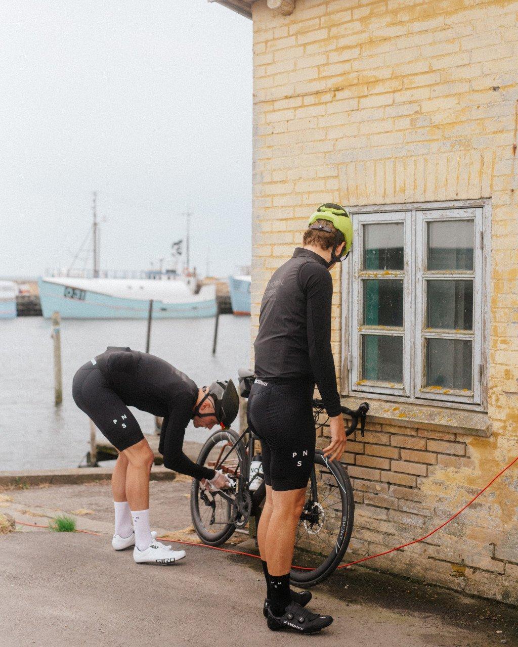 Two cyclists with bike by brick building near harbor, wearing black cycling kit with branding