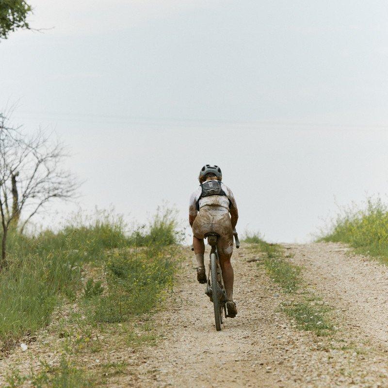 Model cycling on gravel road through countryside wearing tan jersey and shorts with backpack