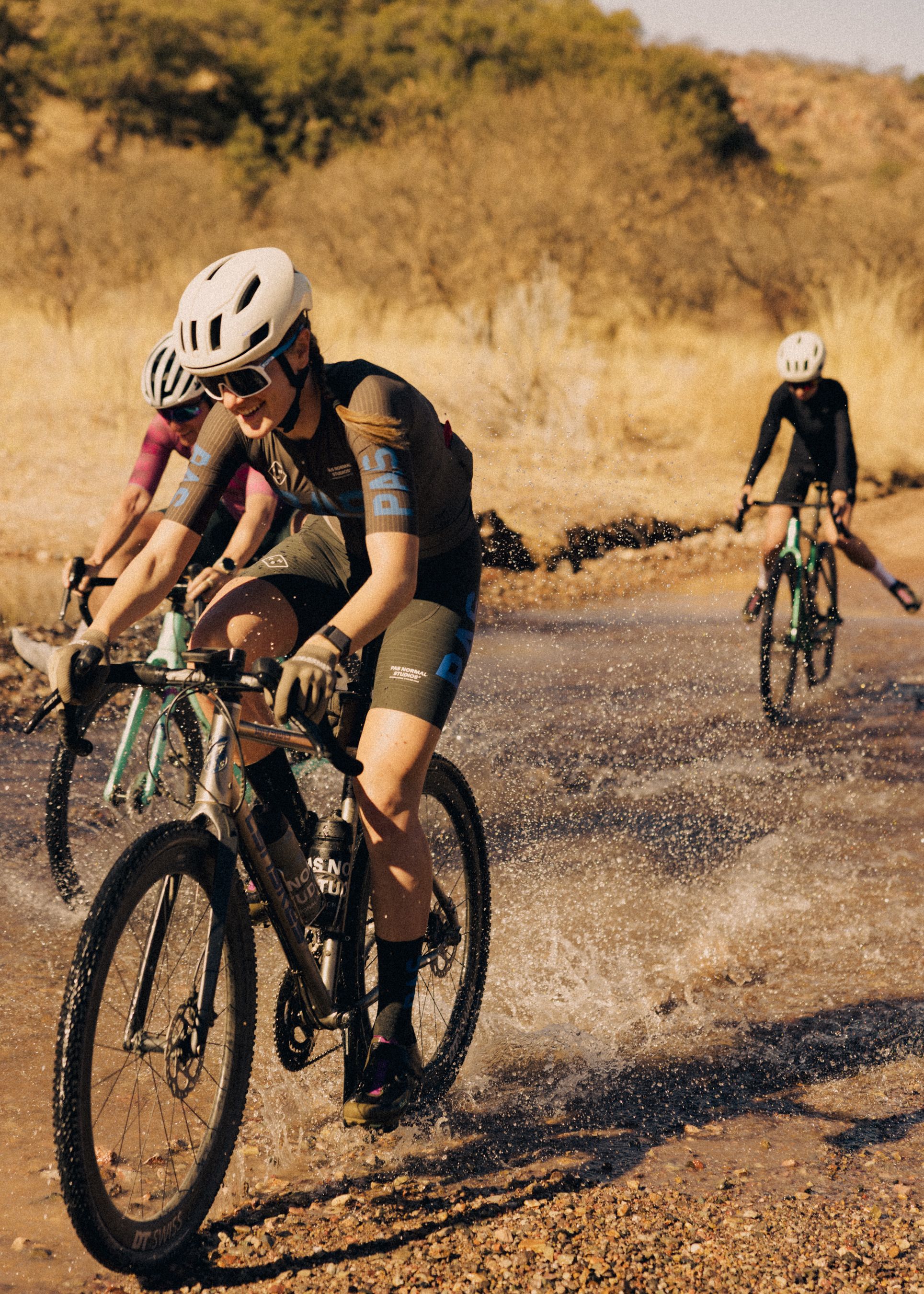 Cyclists wearing black and brown cycling apparel riding through desert terrain with water splashing