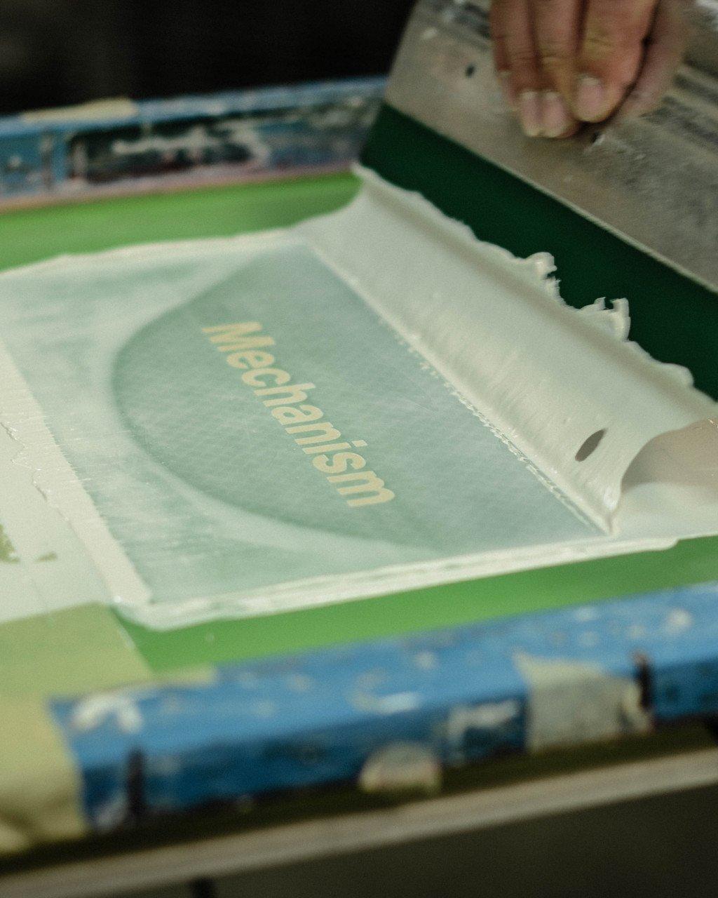 Screen printing process showing Mechanism collection logo being applied to light green fabric