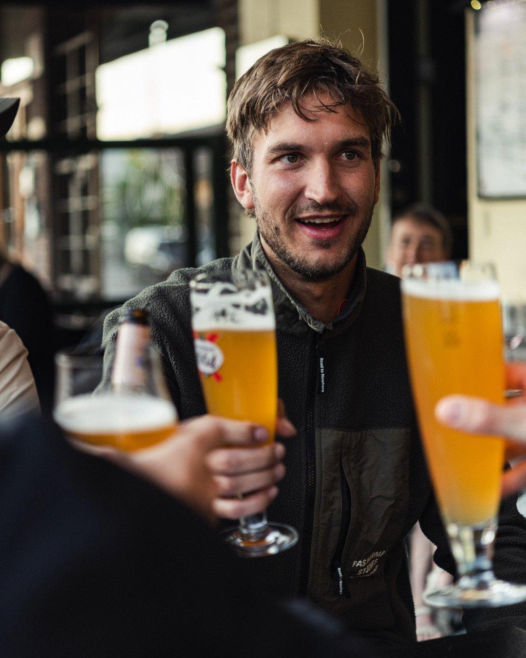 Model wearing dark green zip-up jacket holding beer glass in pub setting