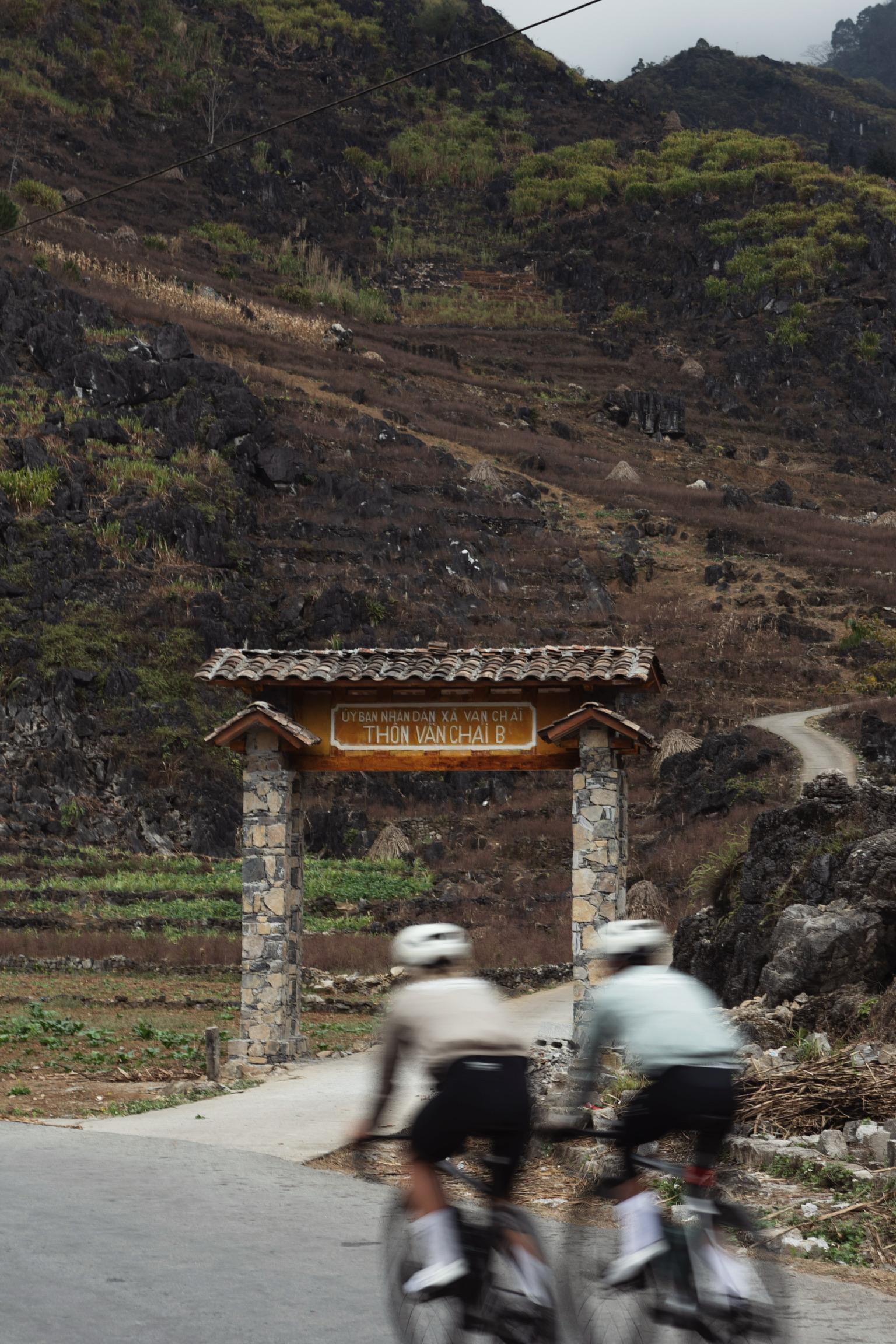 Two cyclists in motion blur passing under stone village entrance gate in mountainous Vietnamese terrain