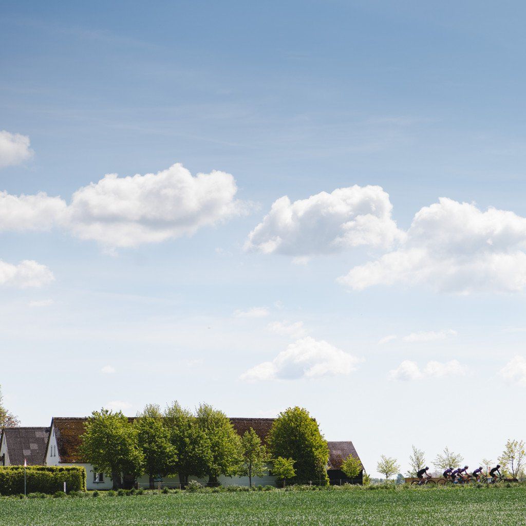Group of cyclists riding through rural countryside with traditional houses and green fields under cloudy sky