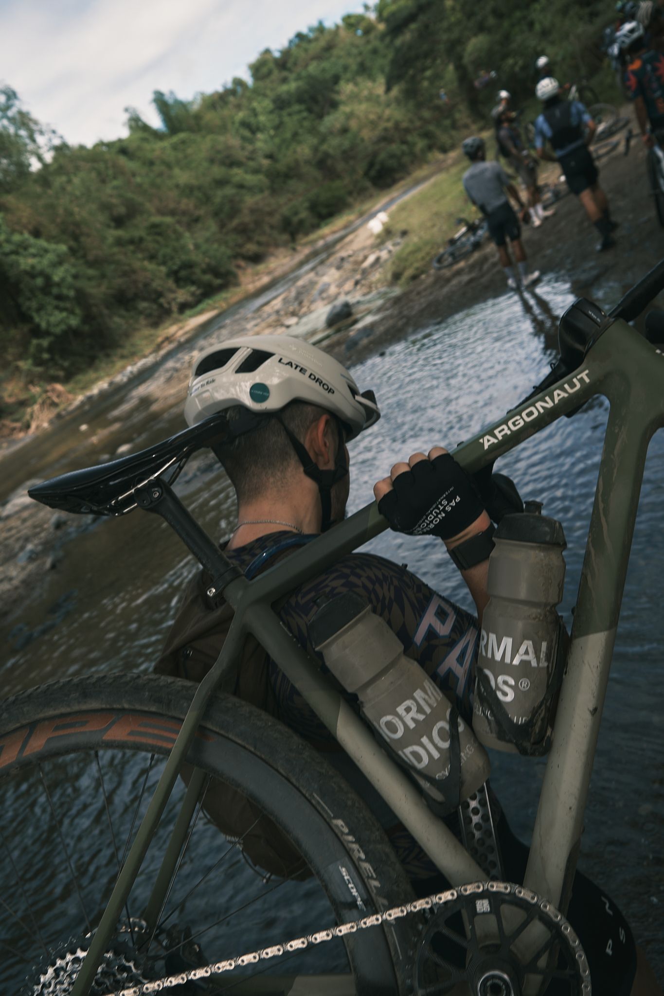 Cyclist with gravel bike by water crossing, wearing white helmet and dark cycling gloves with logo branding