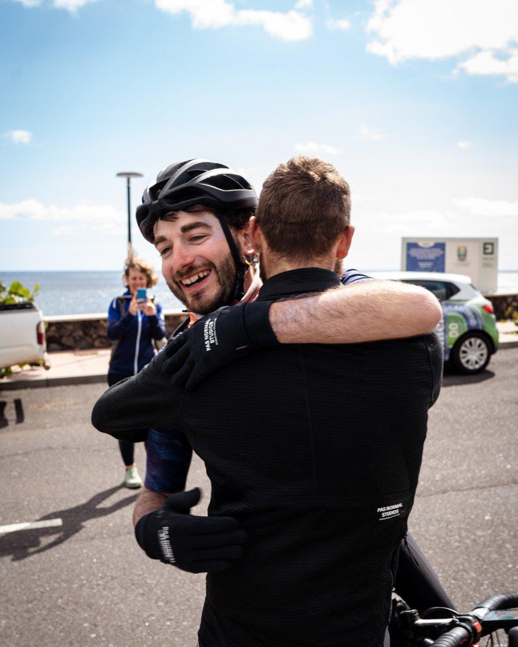Two cyclists embracing after completing a ride, wearing black cycling apparel with helmet